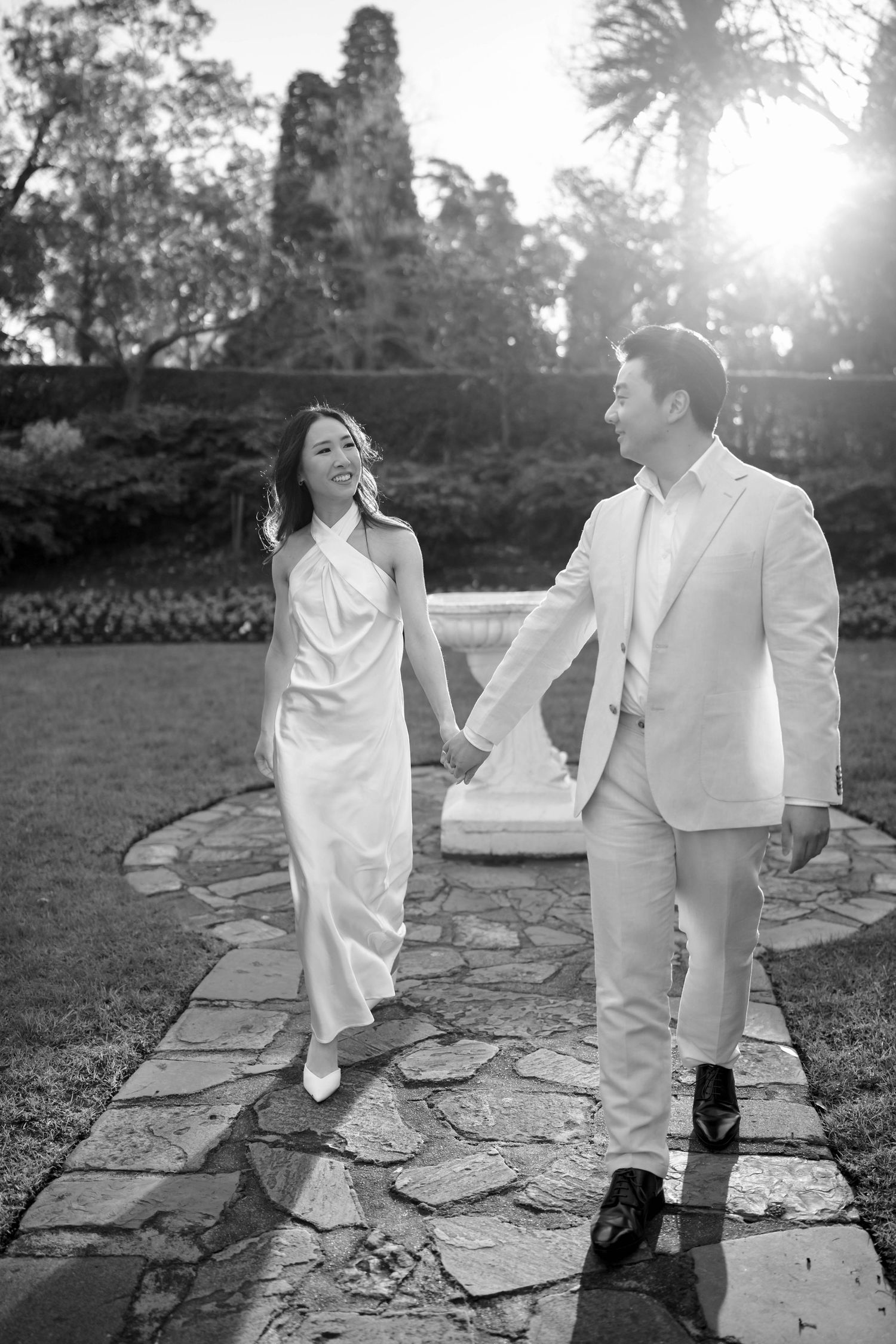 Black and white photo of a smiling couple in wedding attire walking hand in hand along a stone path in a garden.