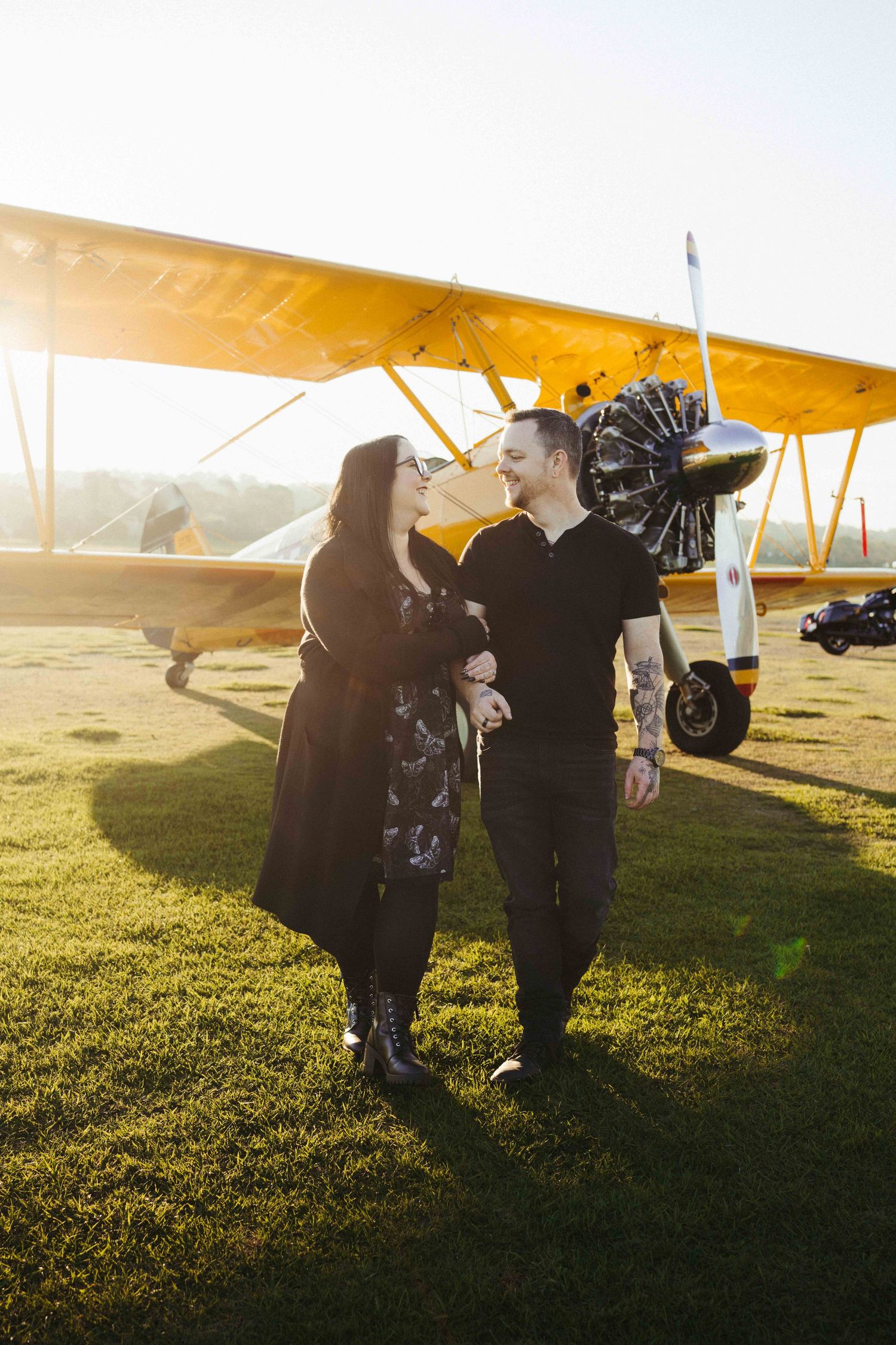Couple sharing a romantic moment in front of a vintage yellow biplane on a sunny airfield.