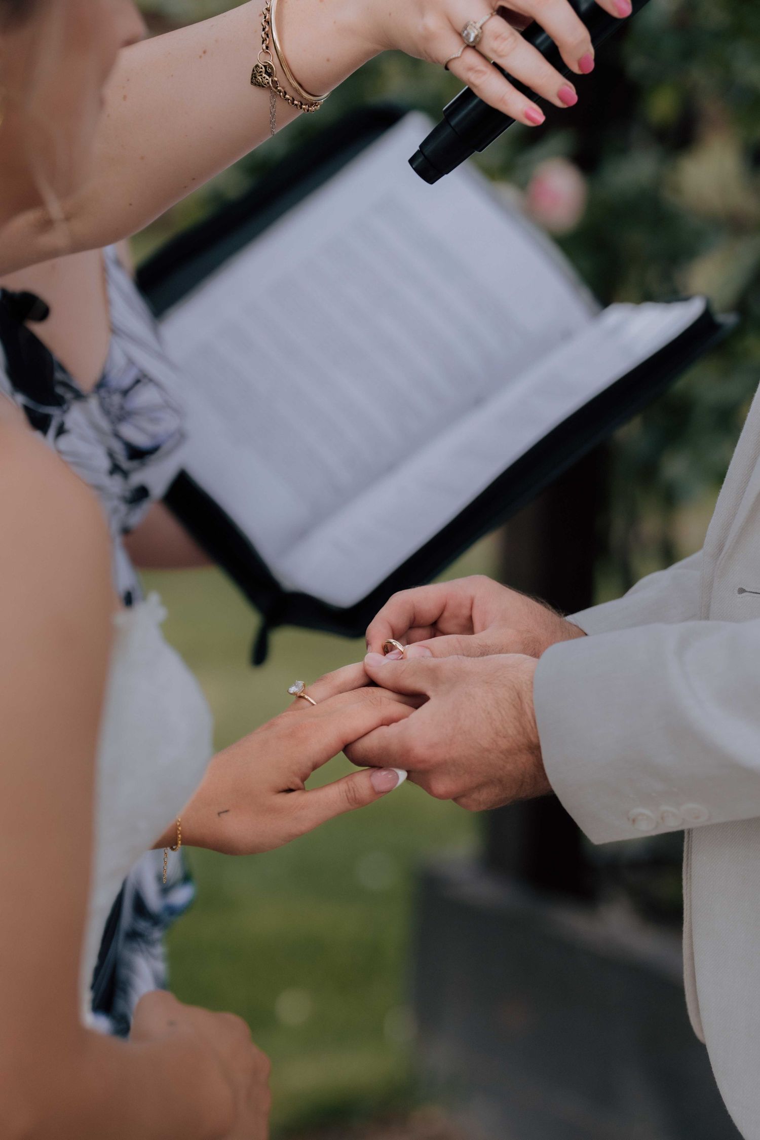 Close-up of groom placing wedding ring on bride's finger during outdoor ceremony with officiant holding microphone.