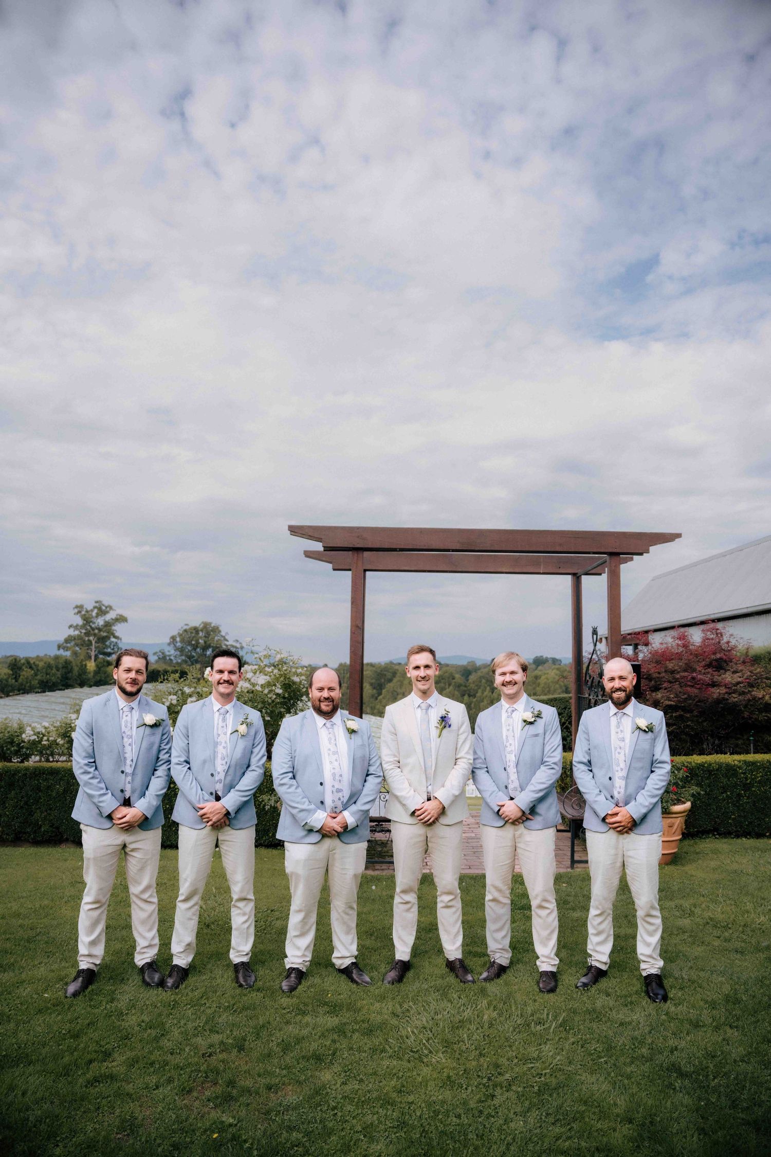Six groomsmen in light blue blazers and cream trousers posing on lawn under cloudy sky.