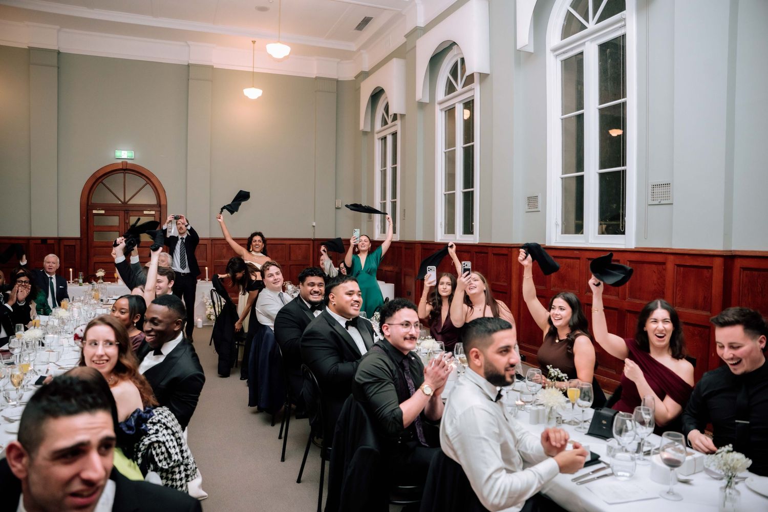 Excited wedding reception guests raise black napkins in celebration inside elegant white ballroom venue.