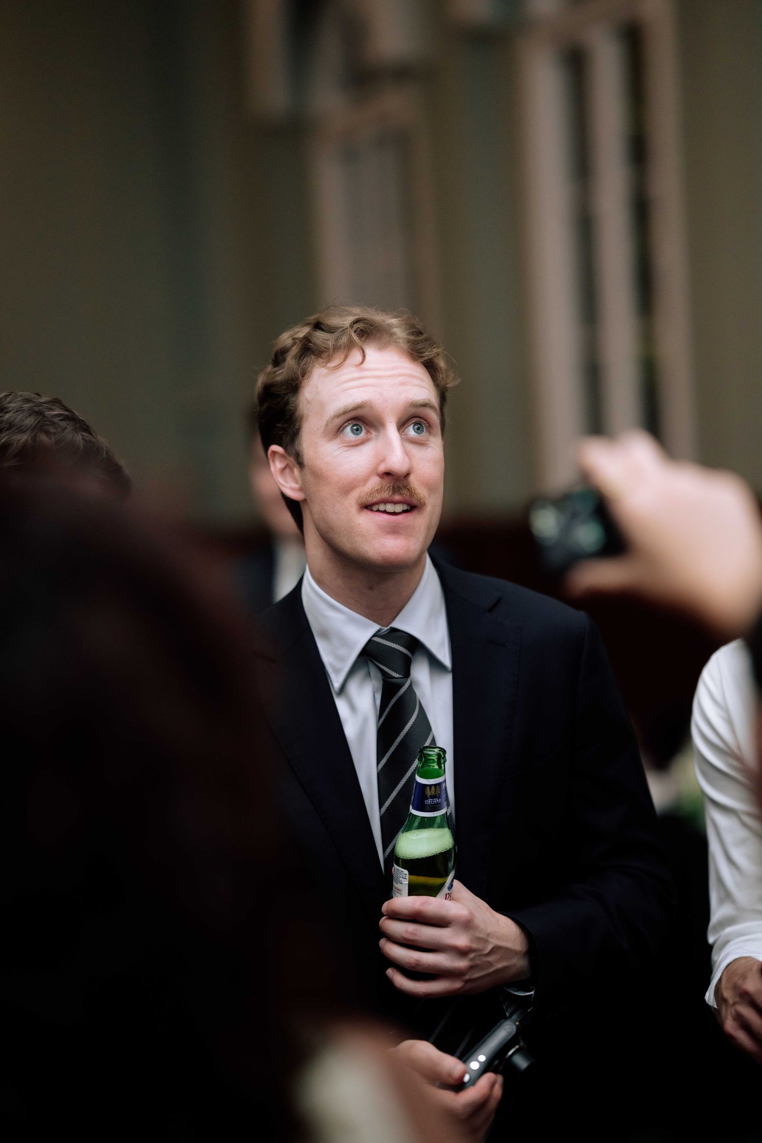 Tall man with mustache in dark suit holding a green bottle at a wedding reception.