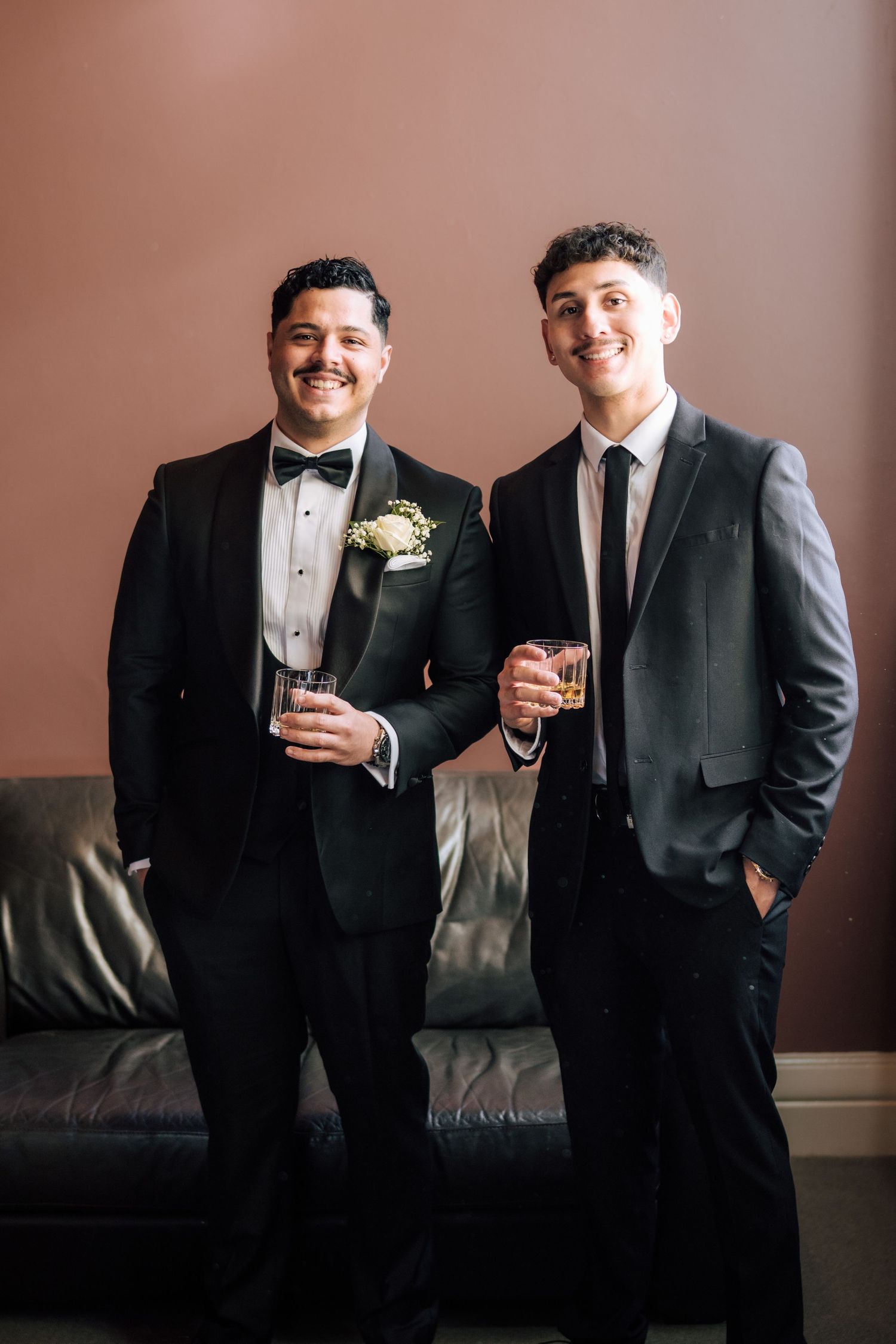 Two grooms in formal suits smile and hold drinks at a wedding celebration.