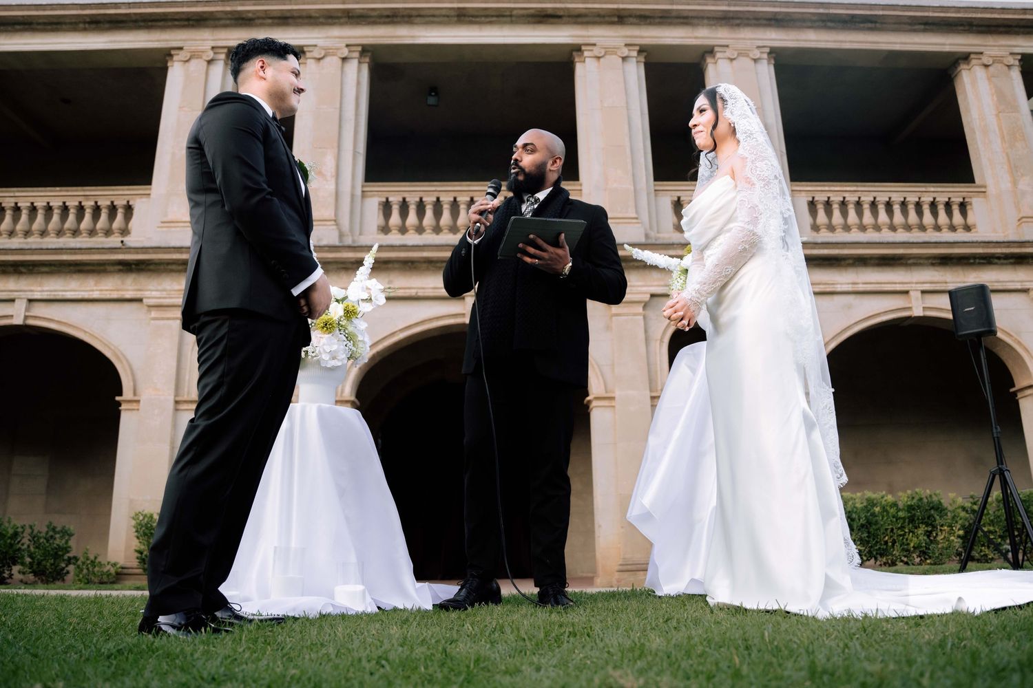Bride and groom exchanging vows with officiant at outdoor ceremony before historic building.