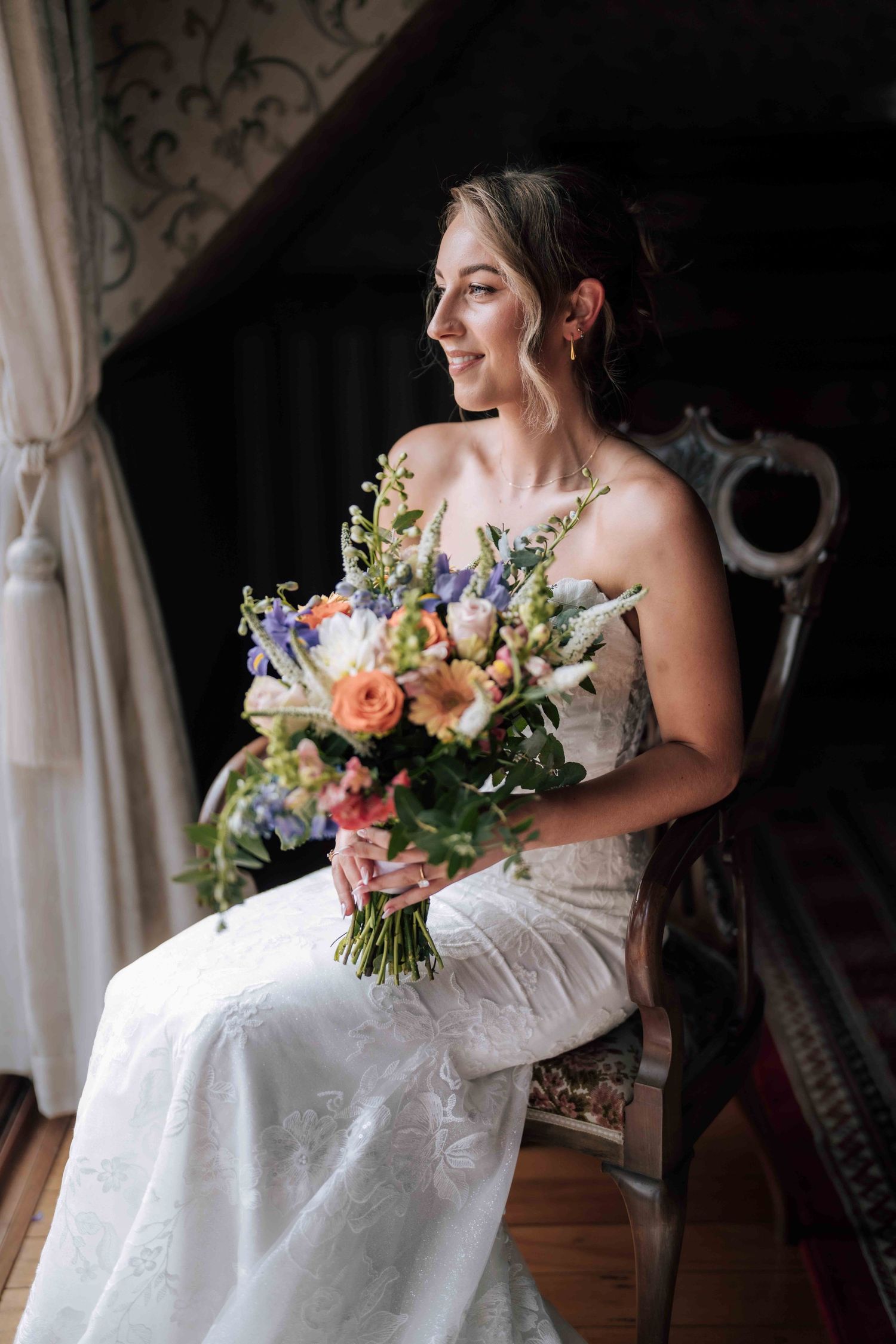 Bride in lace gown sits in vintage chair holding colorful floral bouquet indoors.