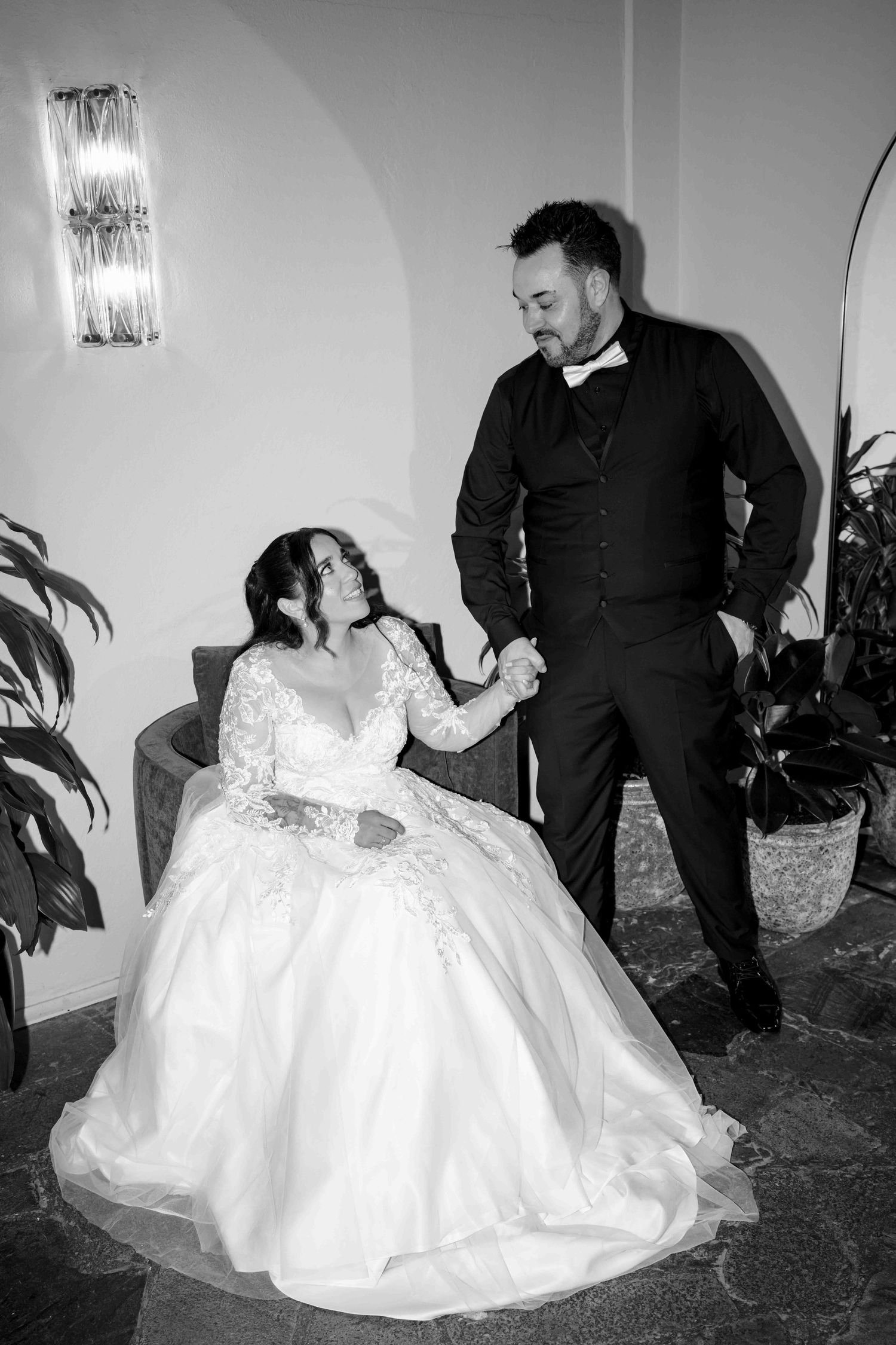 Black and white photo of bride in ball gown sitting while groom in tuxedo holds her hand.