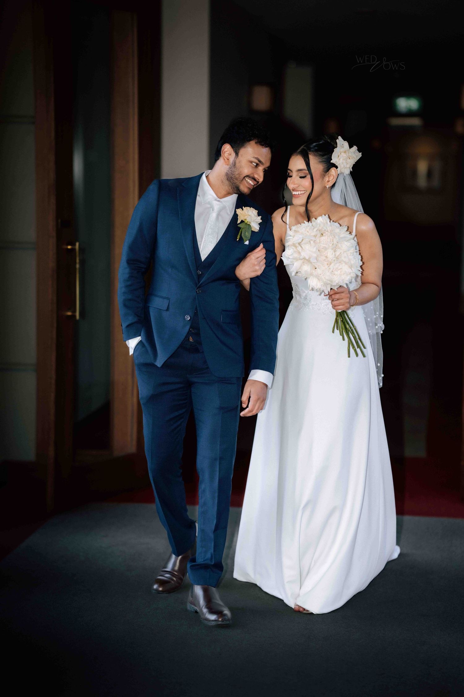 Elegant bride and groom walking together, she holds white bouquet, he wears a navy three-piece suit.