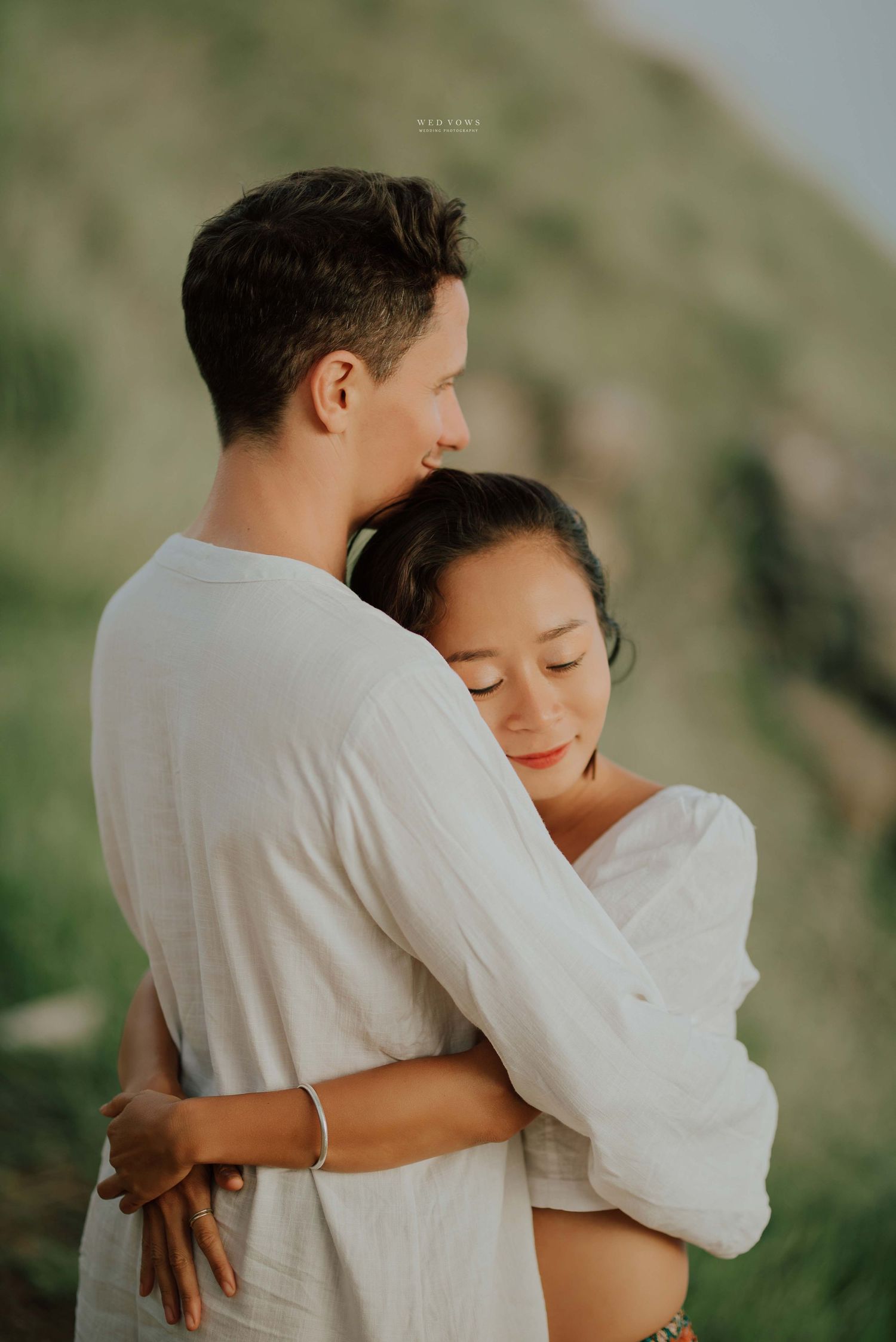 Couple in white outfits sharing a tender embrace with green hills behind them.