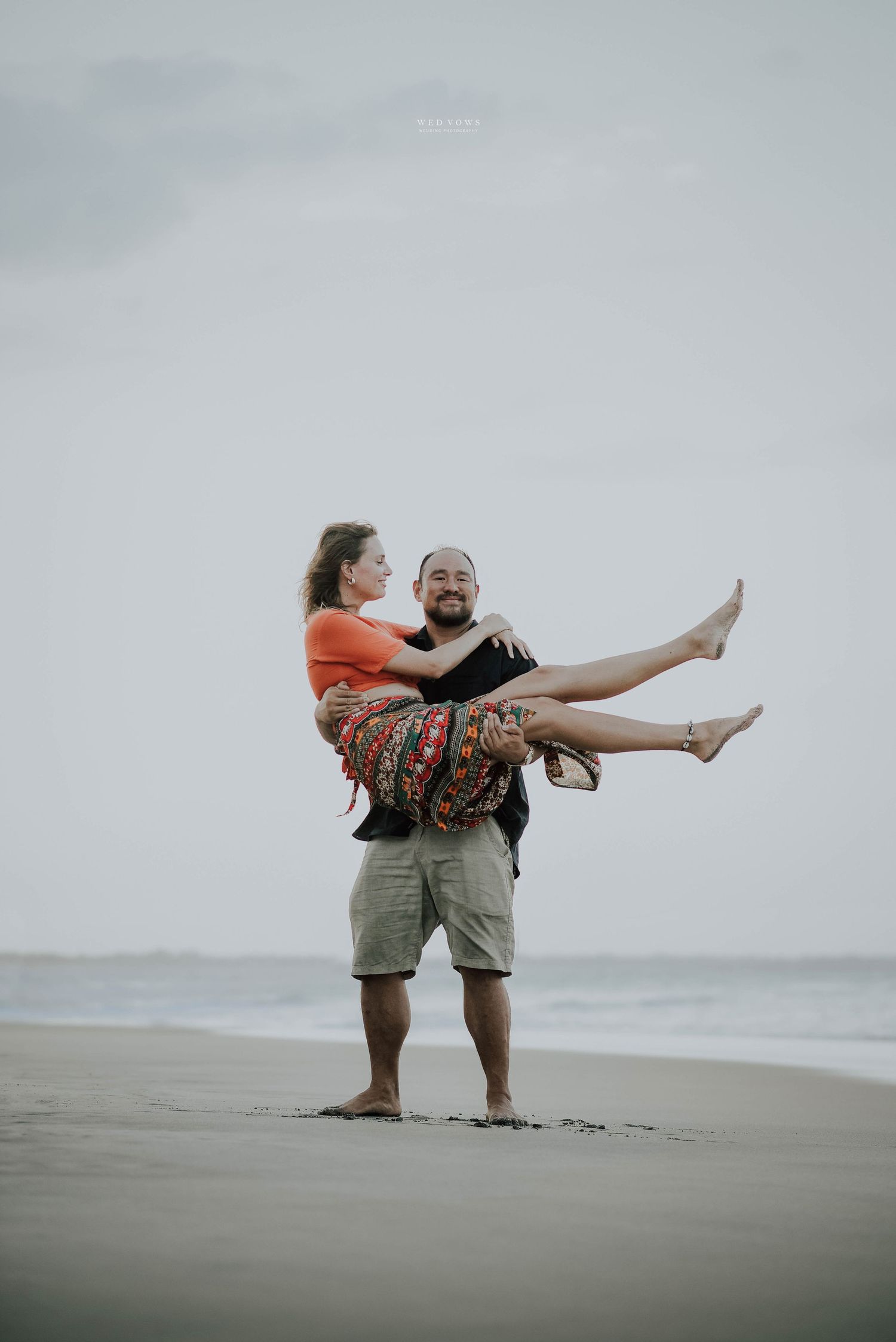 Man carrying woman in his arms on a sandy beach, both laughing joyfully with ocean behind them.