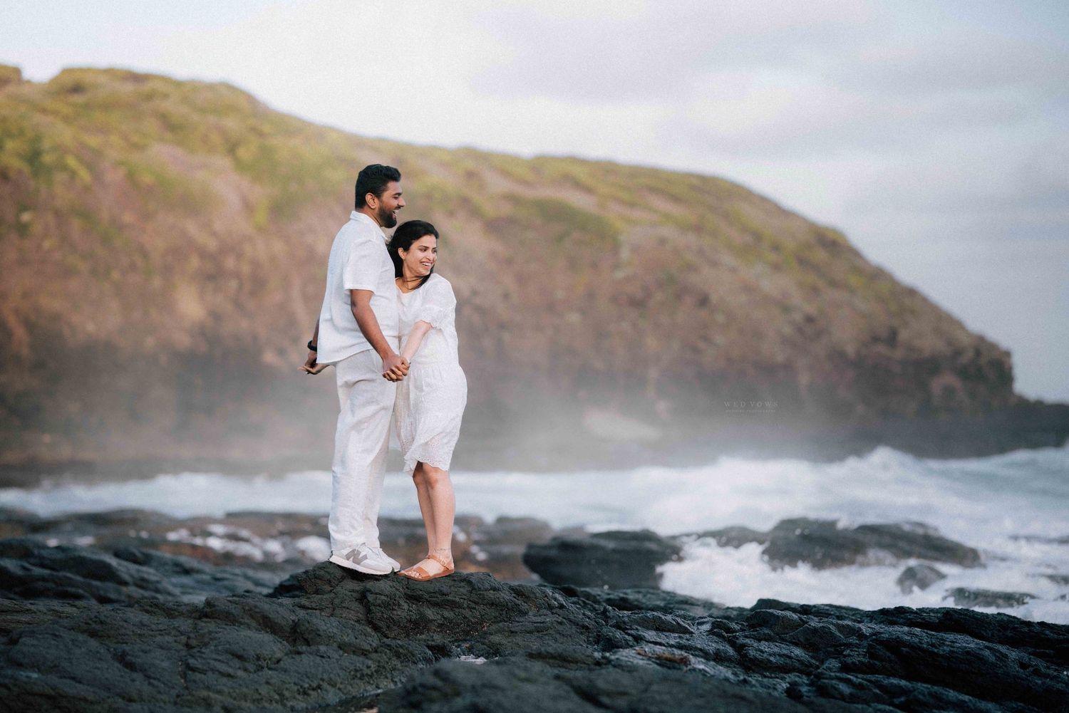 Couple in white embracing on dark rocks with crashing waves and green cliffs behind.