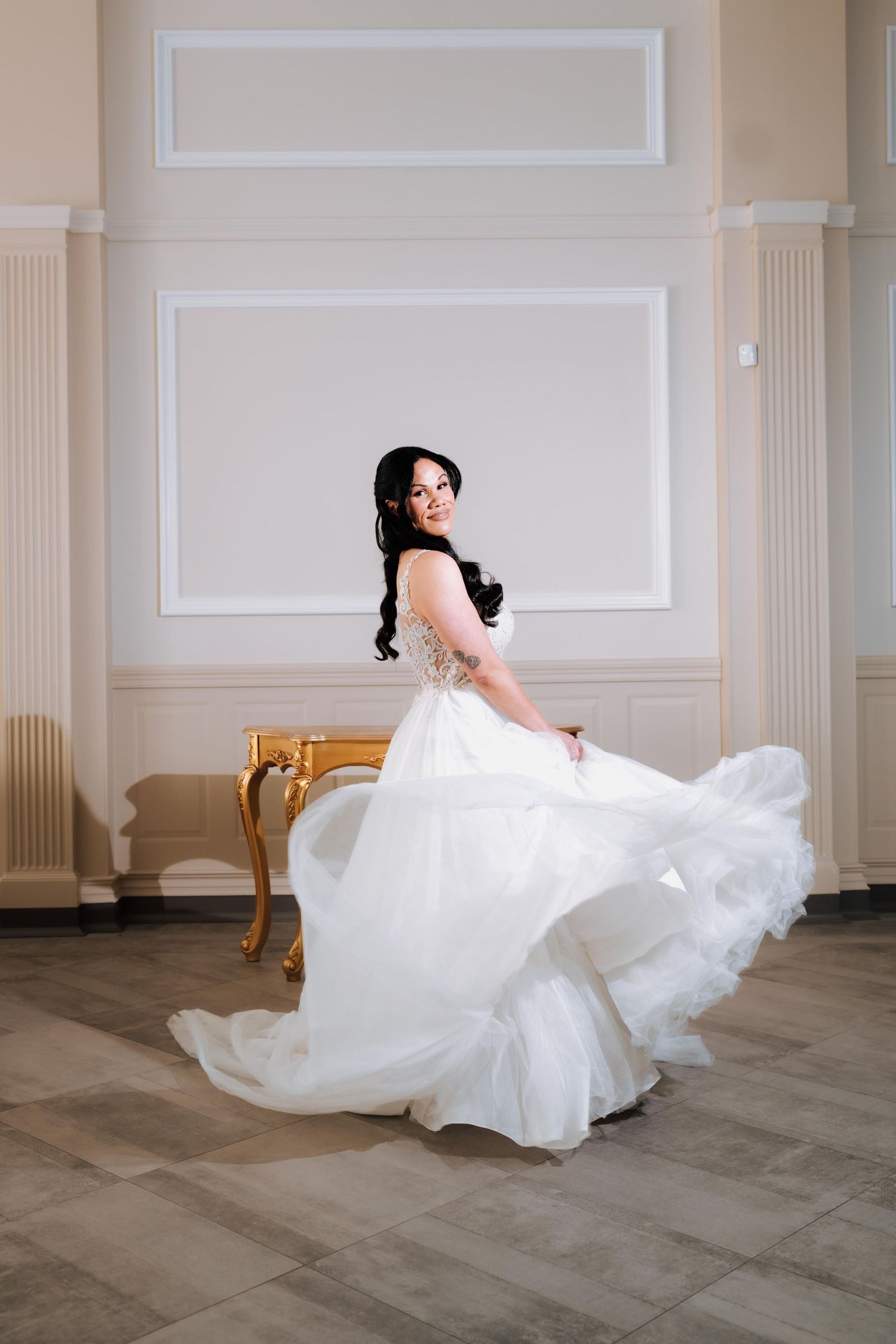 Bride twirling in a flowing white wedding gown in an elegant ballroom with high ceilings.