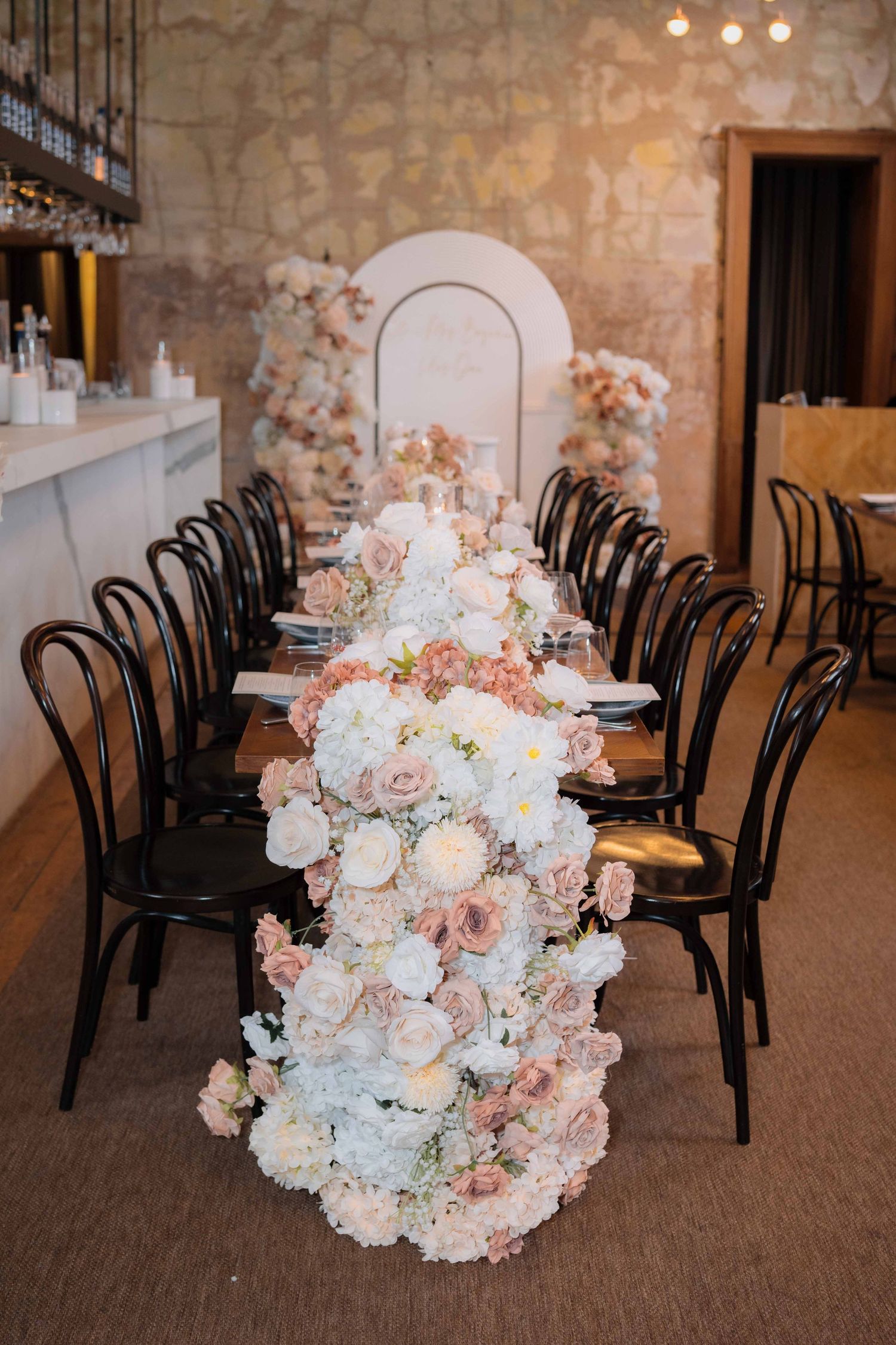 Luxurious wedding reception long table adorned with cascading white and blush pink rose floral runner and black bentwood chairs.