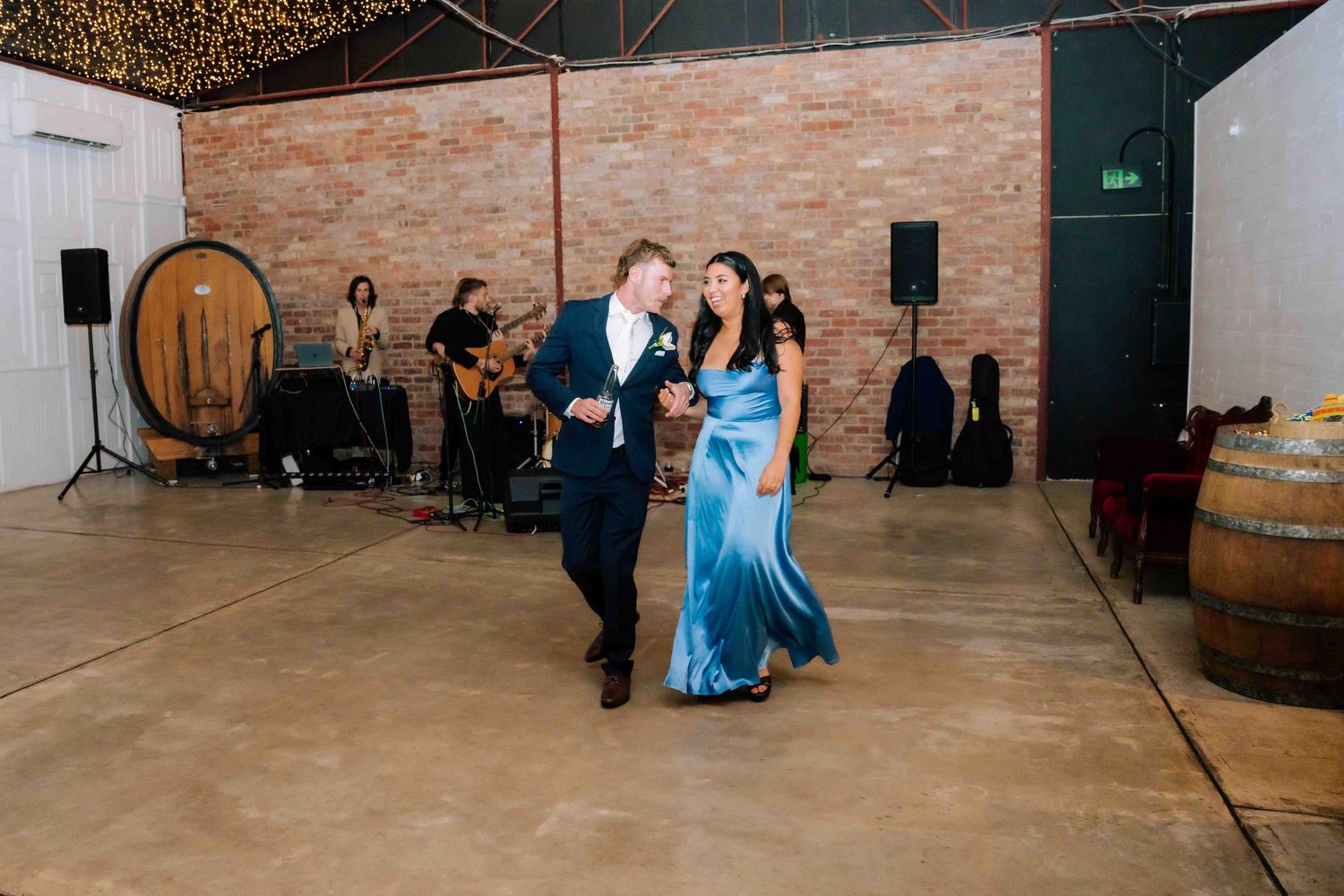 Bridesmaid in blue satin gown dancing with groomsman in navy suit at rustic barn reception.