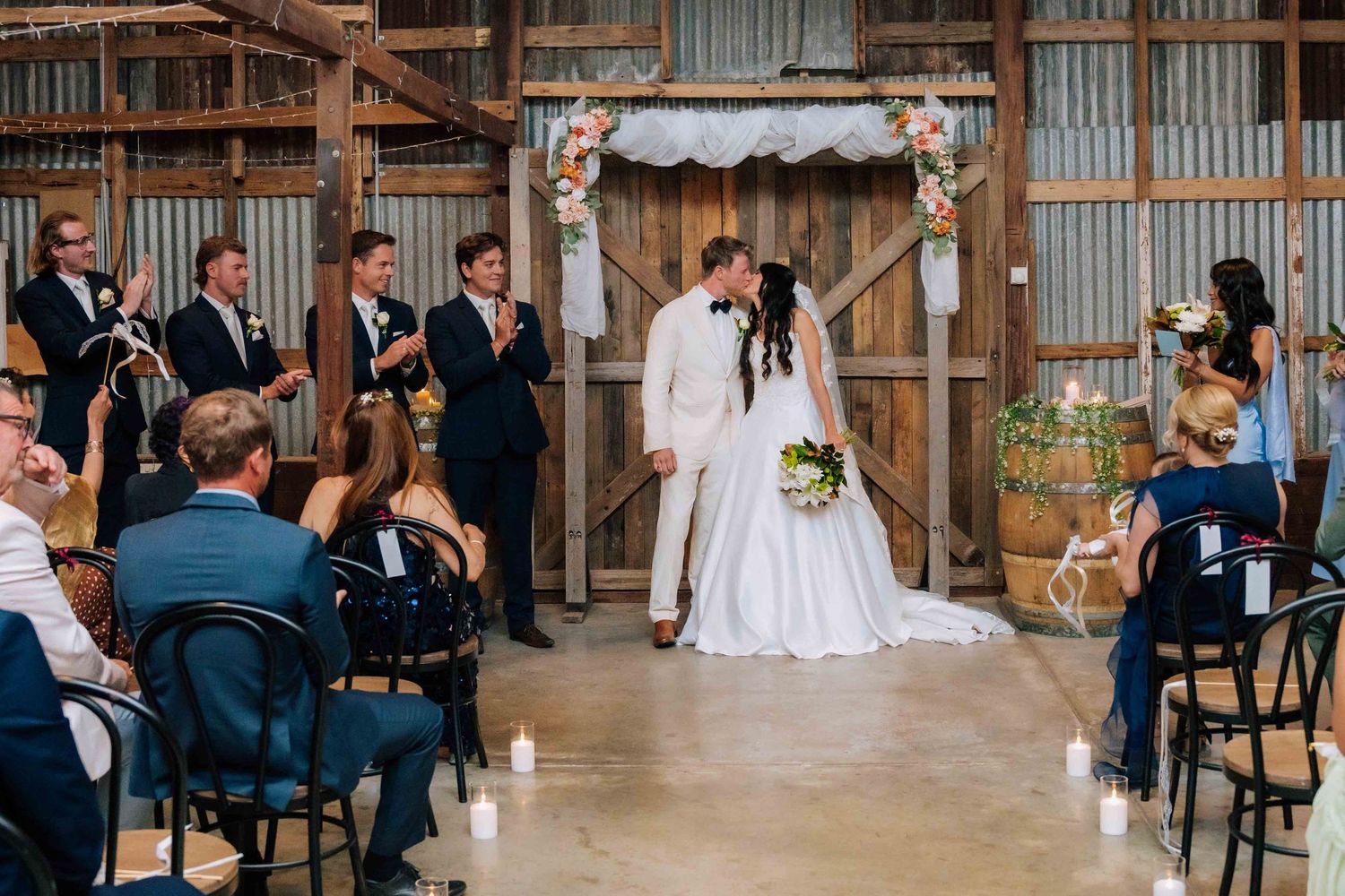 Bride and groom share first kiss at rustic barn wedding ceremony with guests applauding.