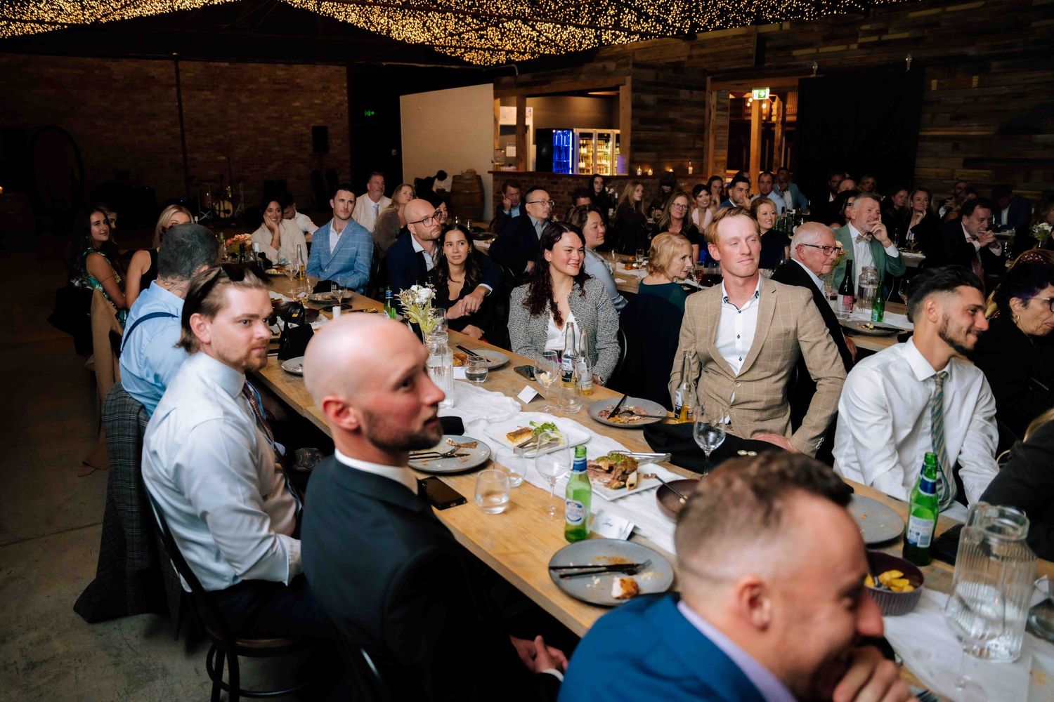 Wedding guests seated at long tables enjoying reception dinner under fairy lights in rustic winery venue.