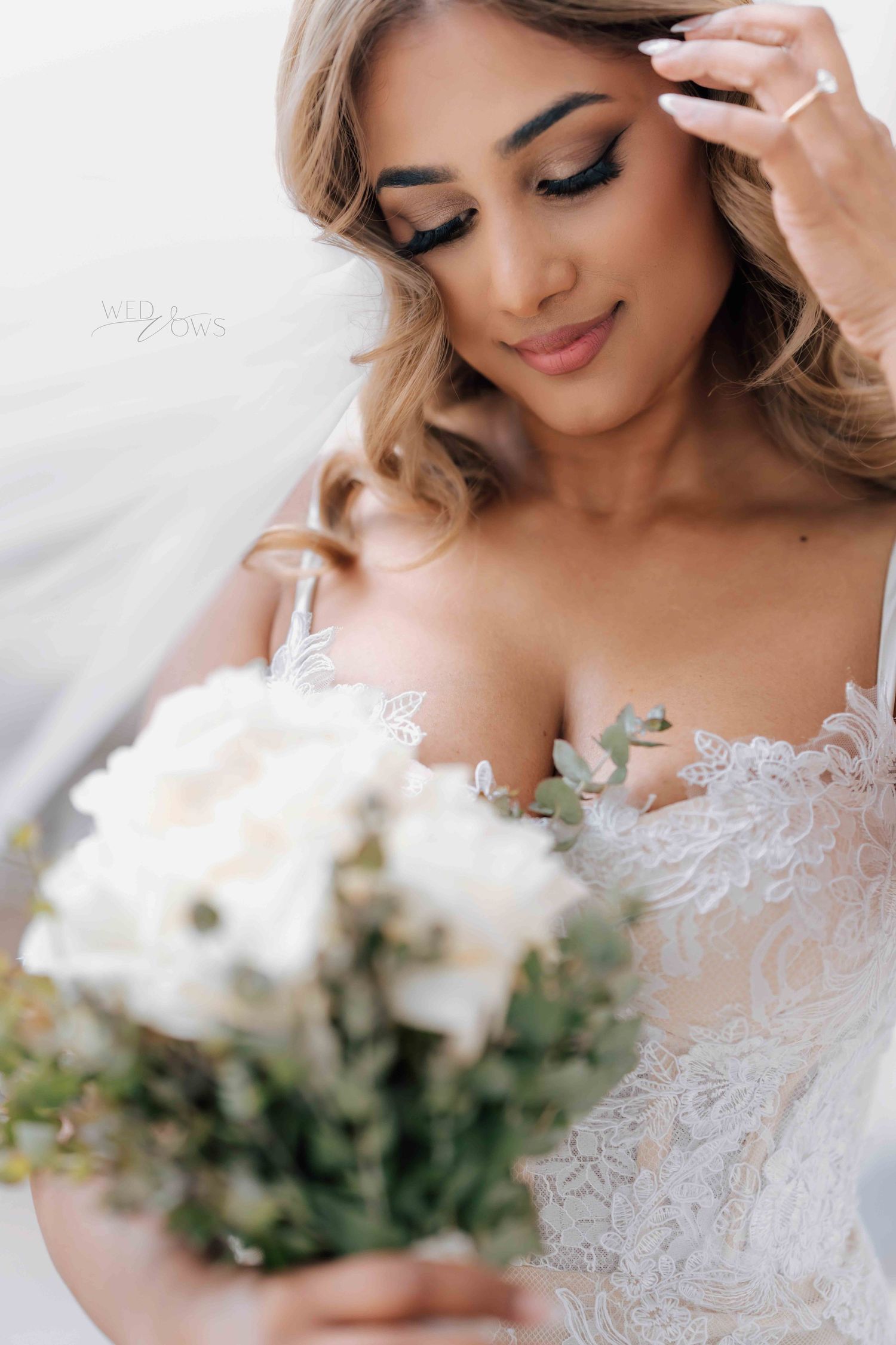 Beautiful bride in lace gown holds white floral bouquet with eyes closed on wedding day.