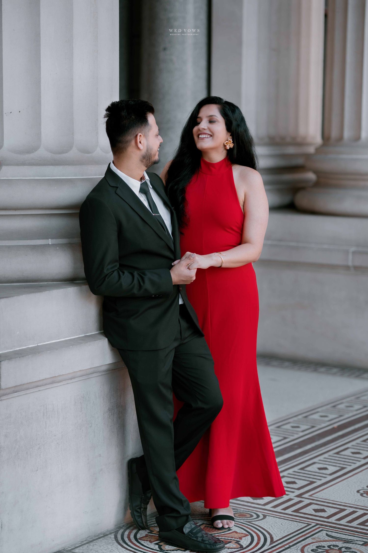 Couple in elegant red gown and dark suit posing by classical columns in a romantic engagement photo.