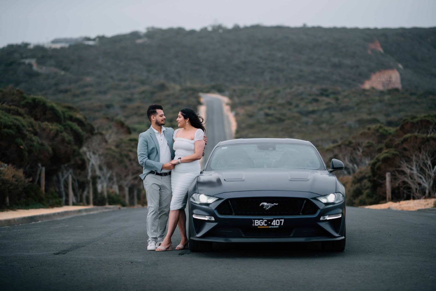 Couple in casual grey outfits stand together beside a dark Ford Mustang on a scenic mountain road.