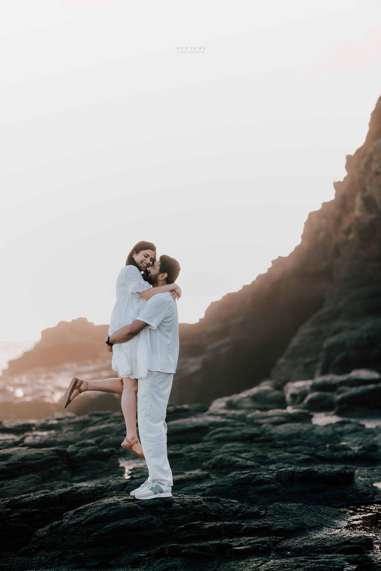 Man lifting woman in white outfits sharing a joyful moment on rocky seaside cliffs at sunset.