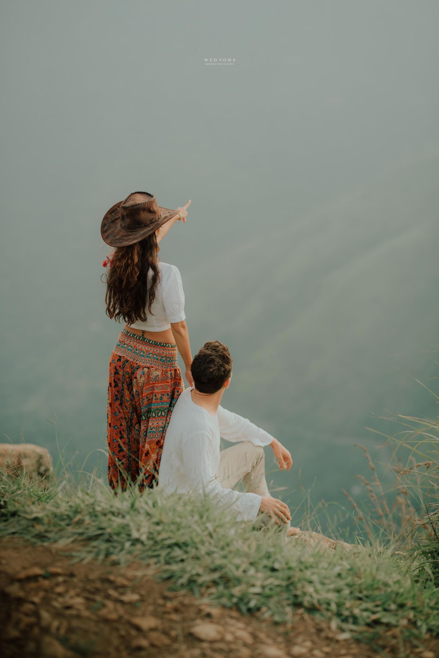 A couple sits on a grassy cliff edge overlooking misty mountains, woman in boho hat and tribal pants points to the distance.