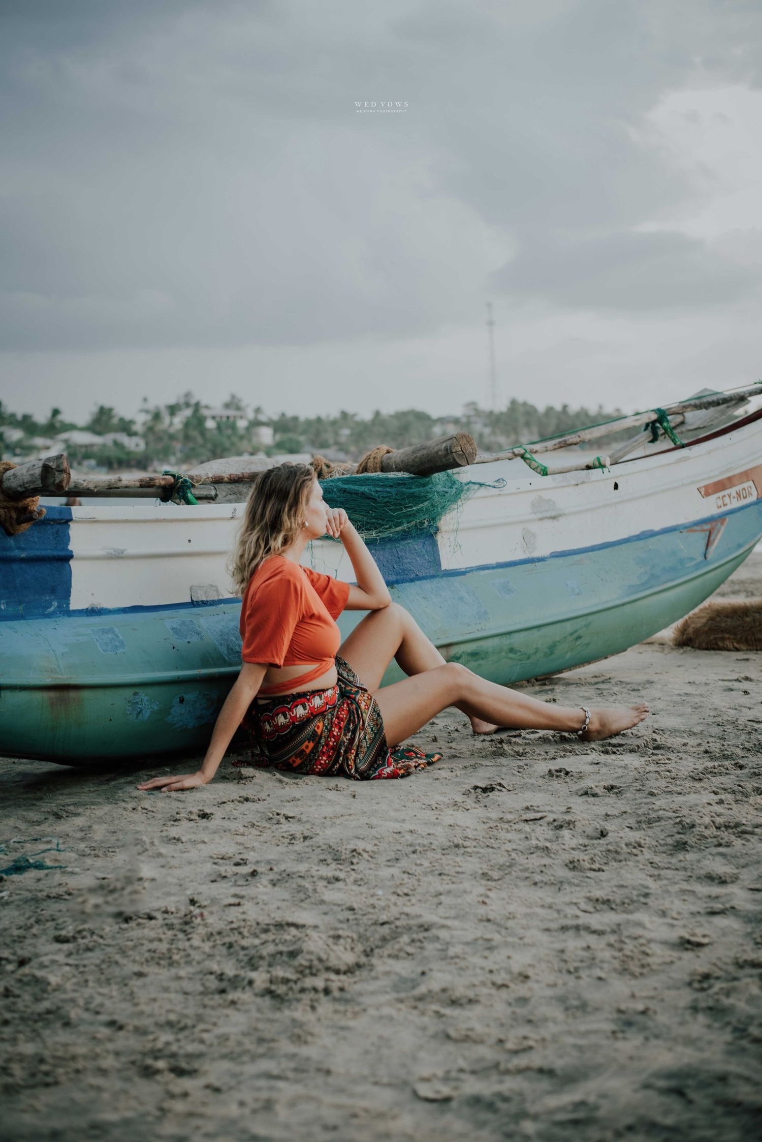 Woman in orange top sitting beside colorful fishing boats on a cloudy beach shore.