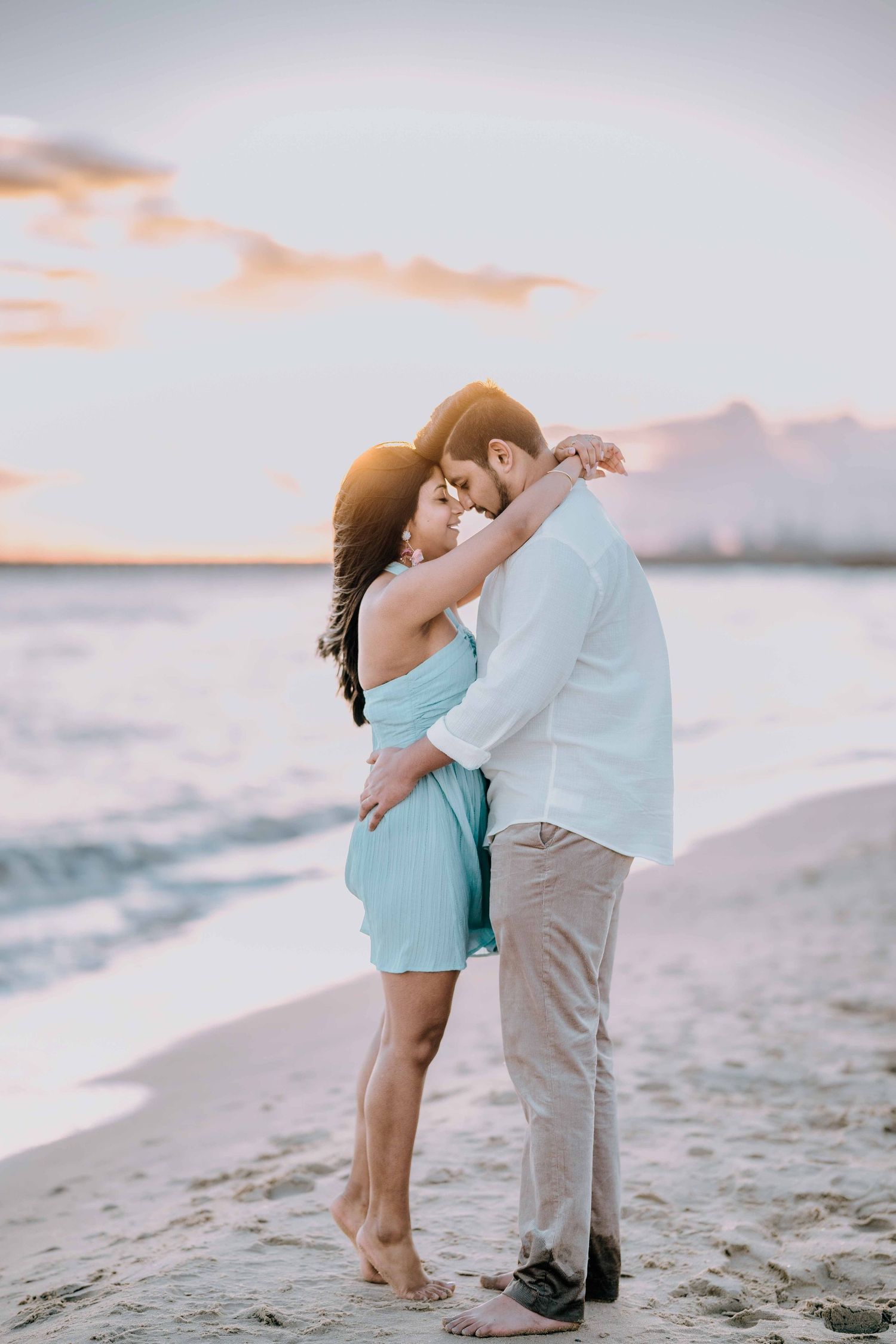 Couple sharing a tender embrace on a beach at golden sunset, woman in light blue dress.