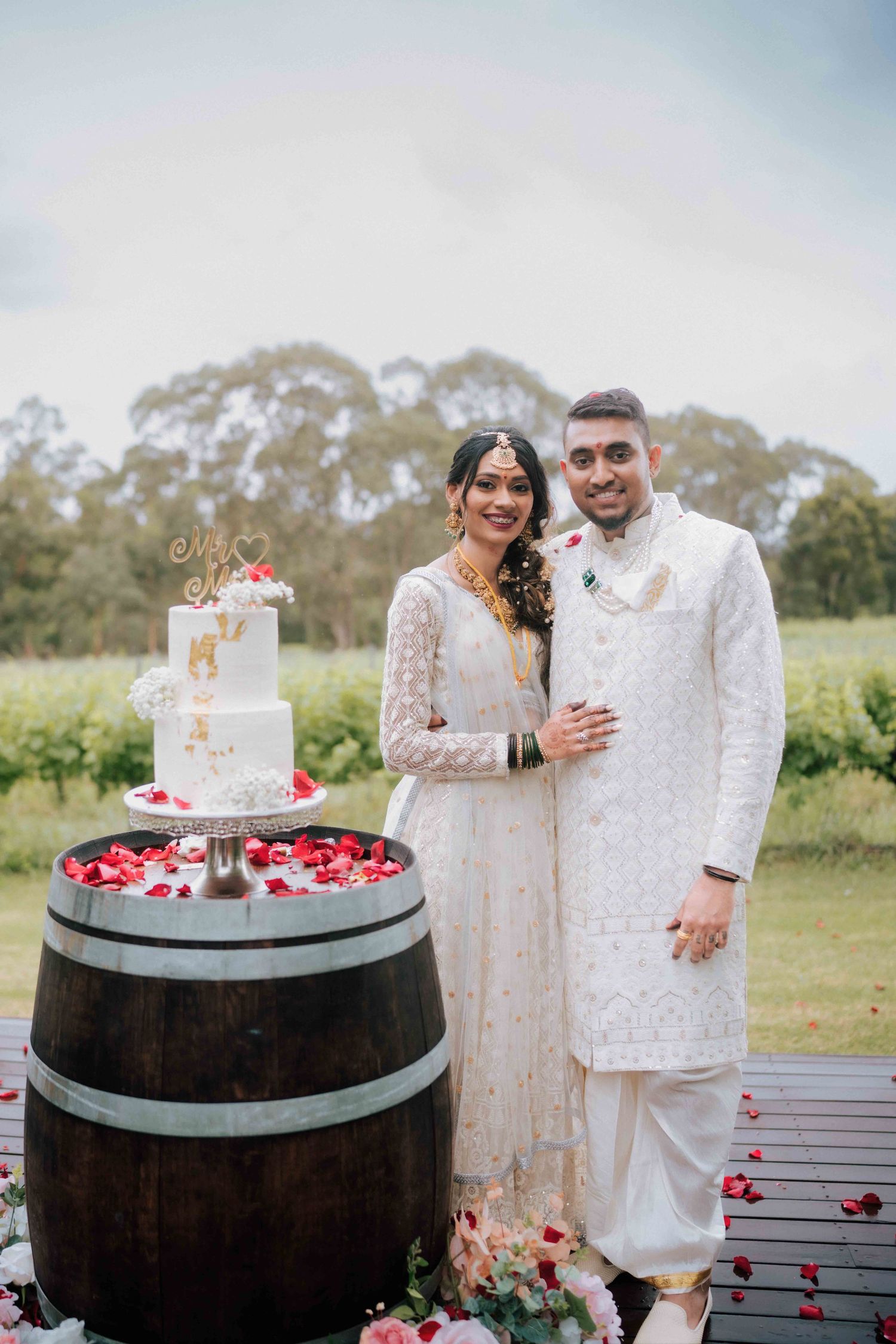 Indian wedding couple in white outfits posing beside tiered wedding cake displayed on a wooden barrel.