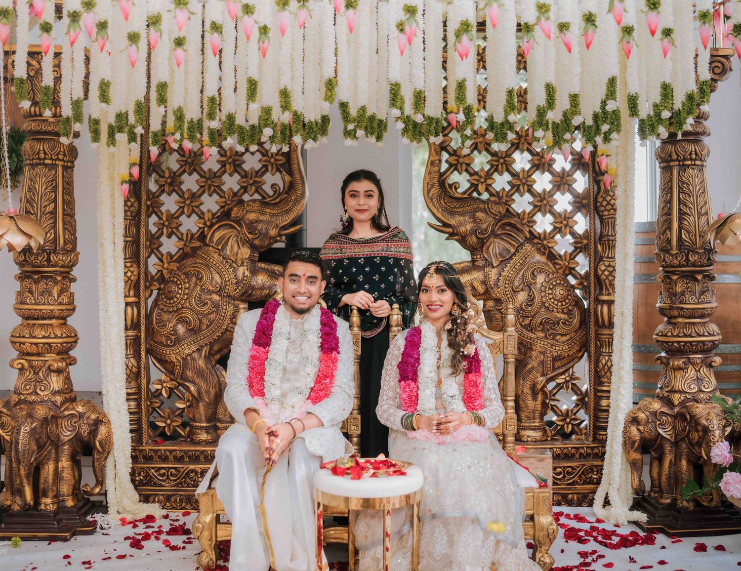 Indian bride and groom seated at ornate mandap with female guest standing behind them smiling.