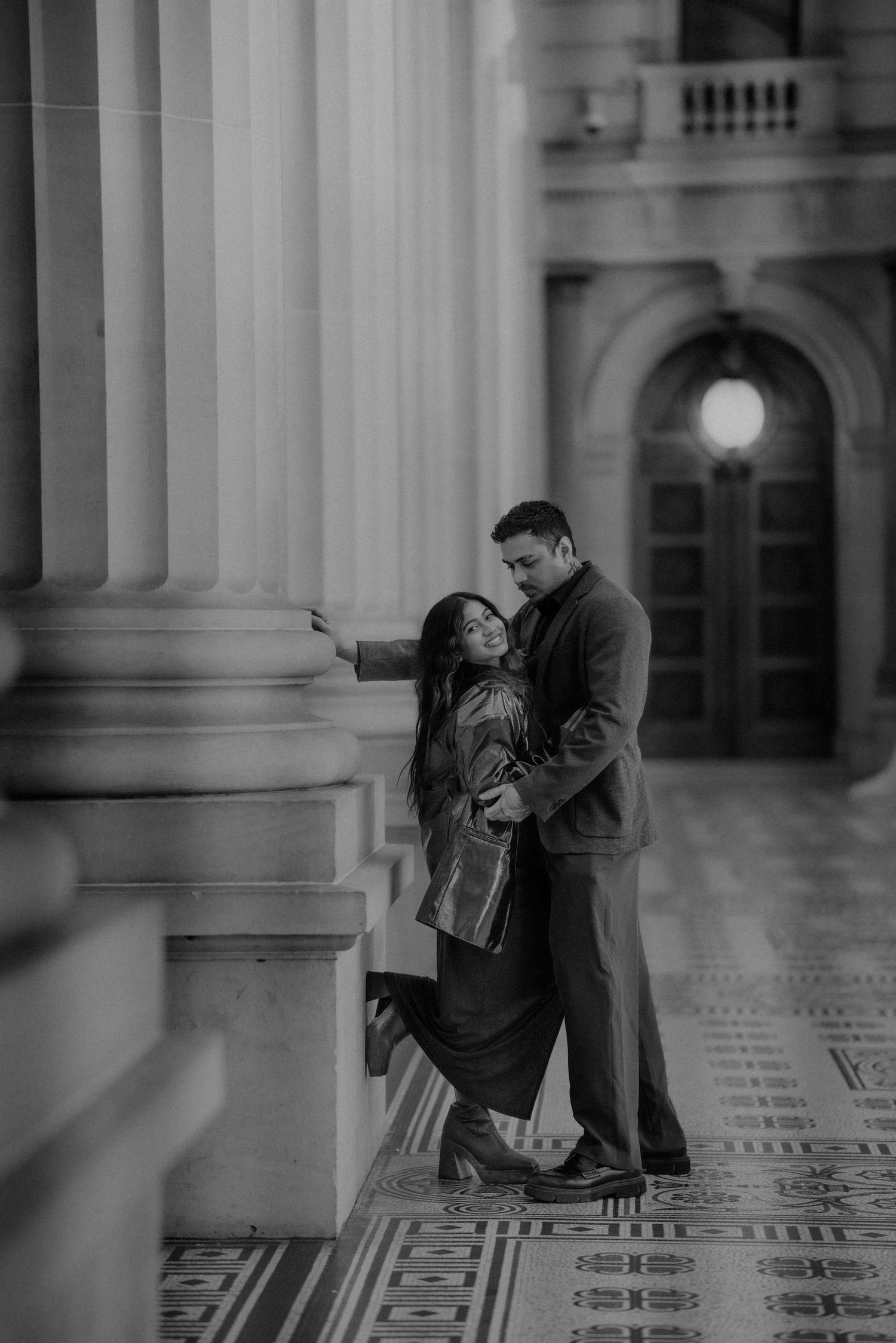 Black and white photo of couple embracing beside grand stone columns in historic building.