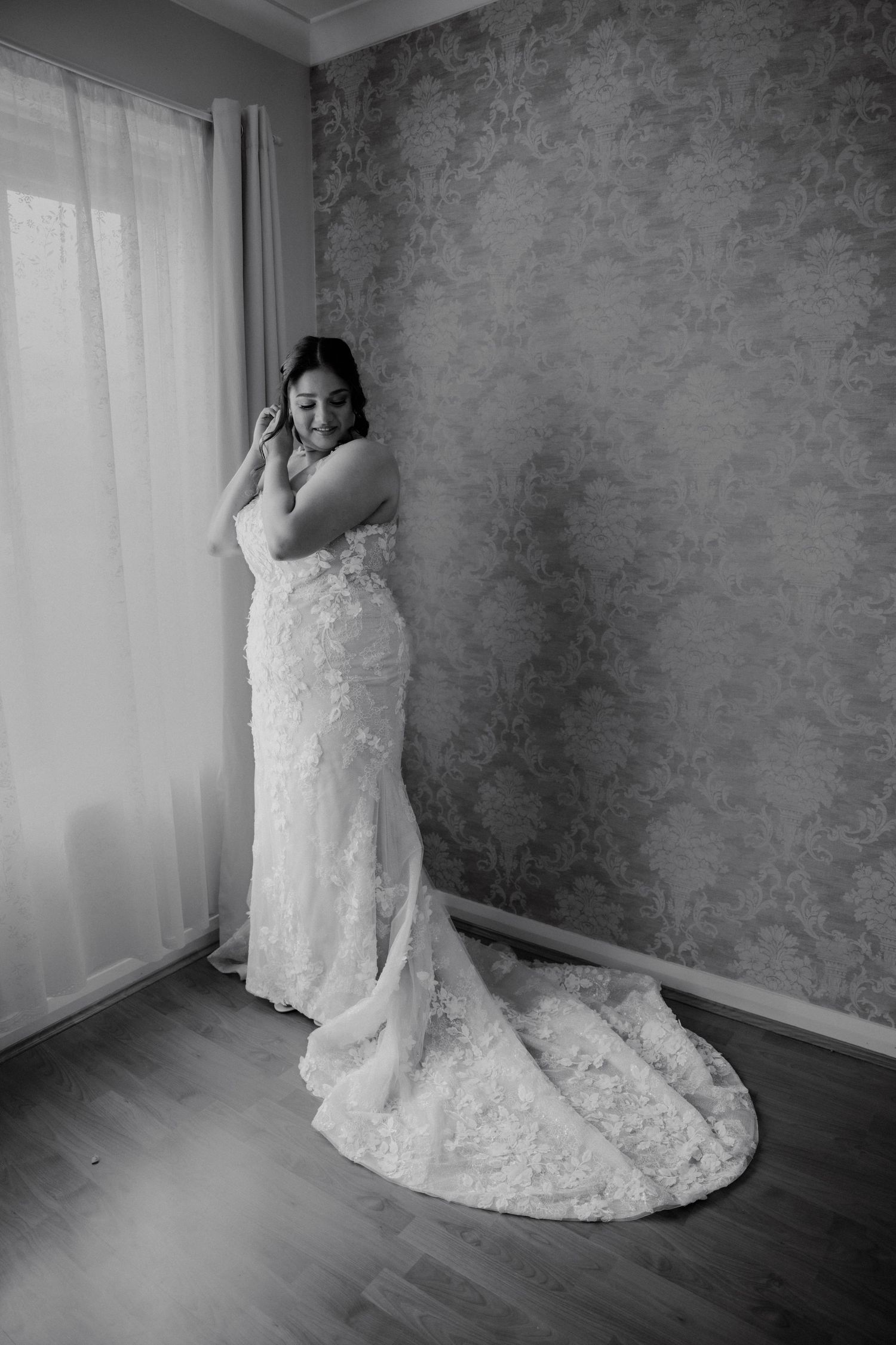 Elegant bride in lace gown with train stands by window adjusting earring in black and white photo.