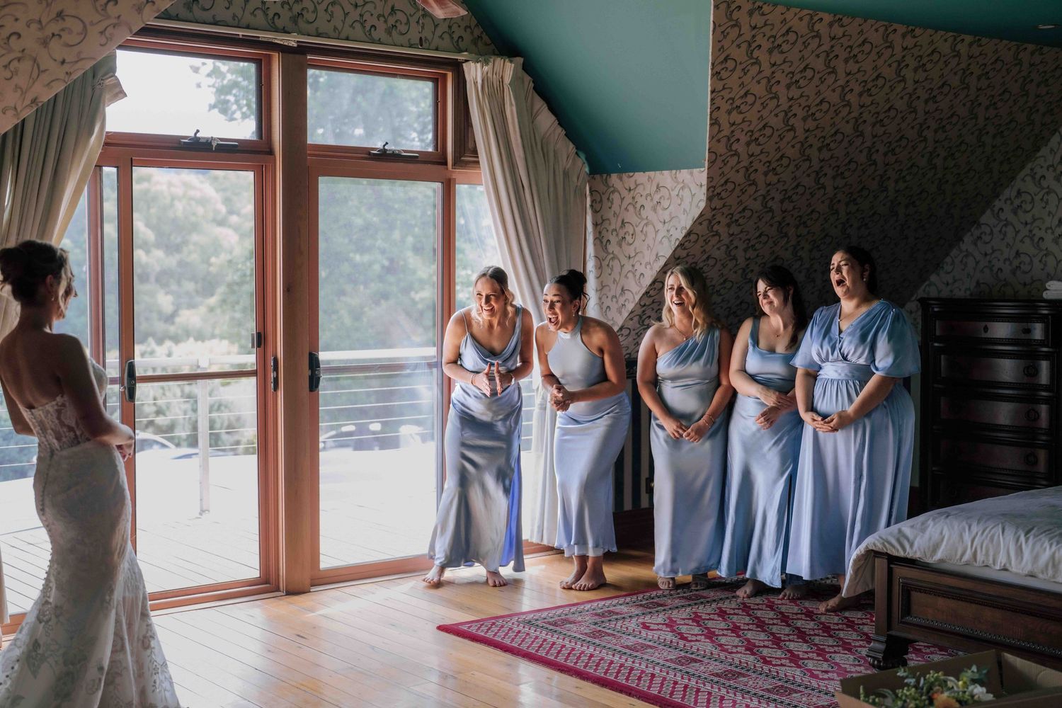 A bride in a white lace gown reveals herself to five bridesmaids in light blue dresses during a first look moment.