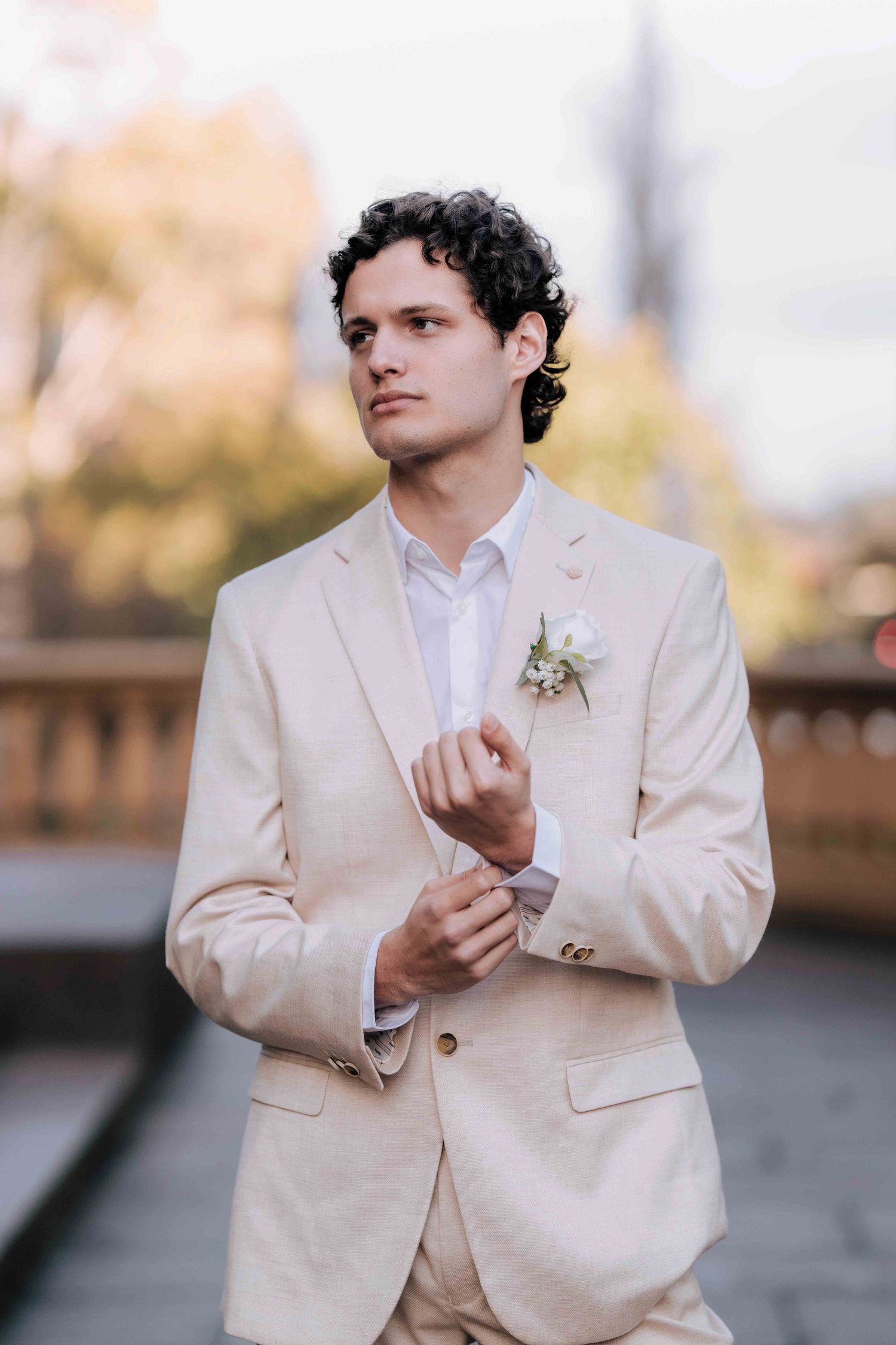 Groom in cream suit adjusting cufflinks with white boutonniere in outdoor wedding portrait.