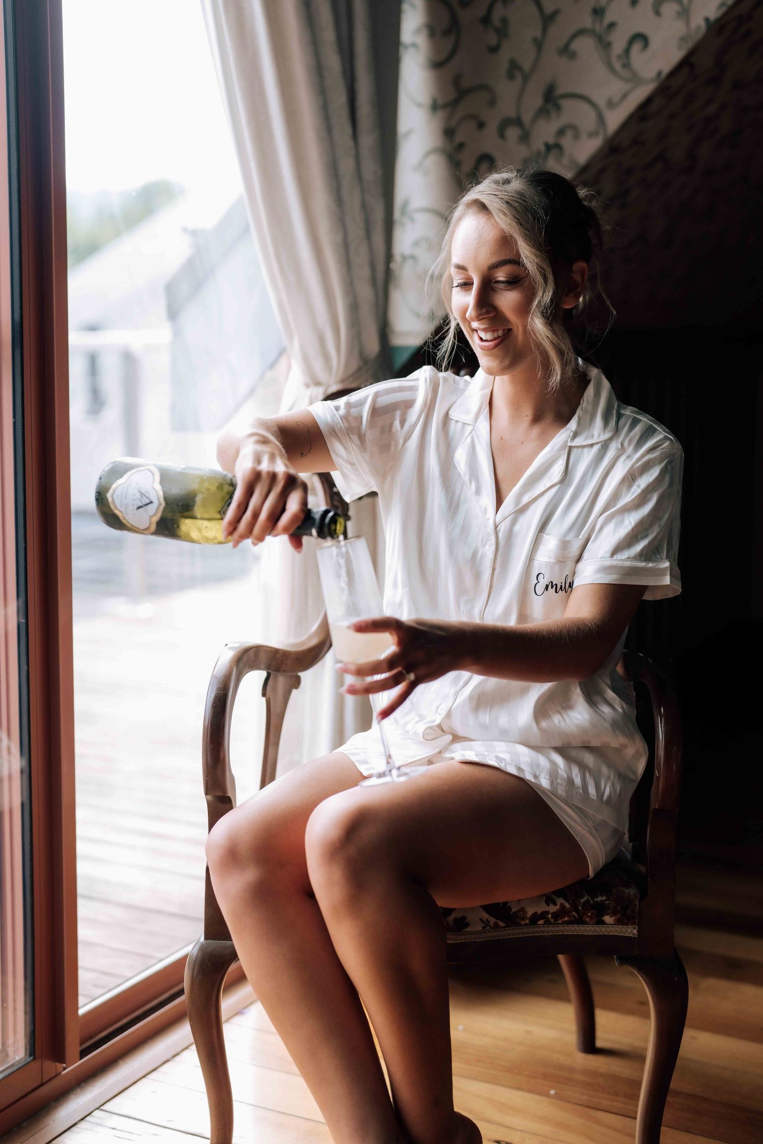 Smiling blonde bride in monogrammed pajamas pouring champagne while seated in an ornate chair by a window.