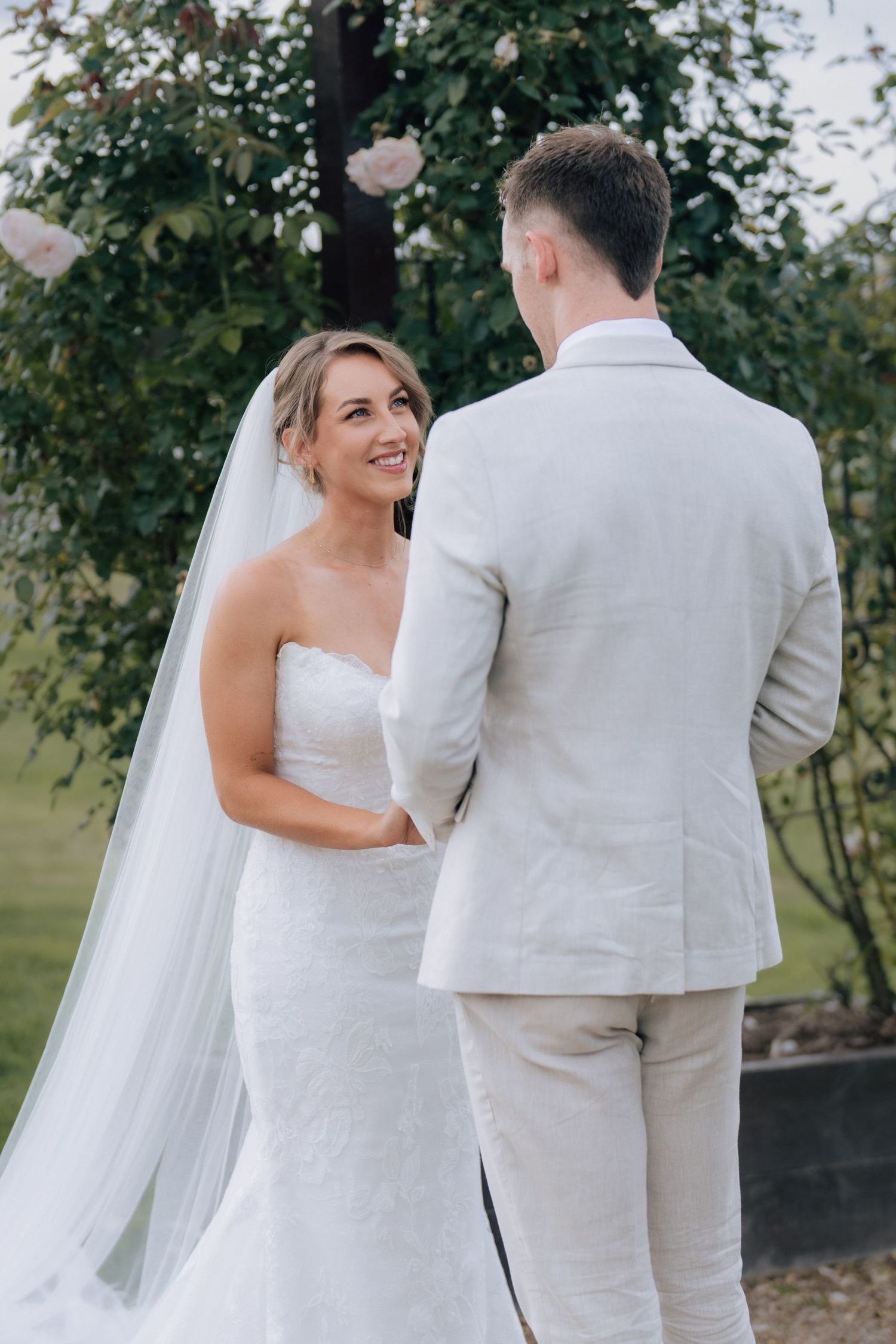 Smiling bride in strapless lace gown holding grooms hands during outdoor wedding ceremony.