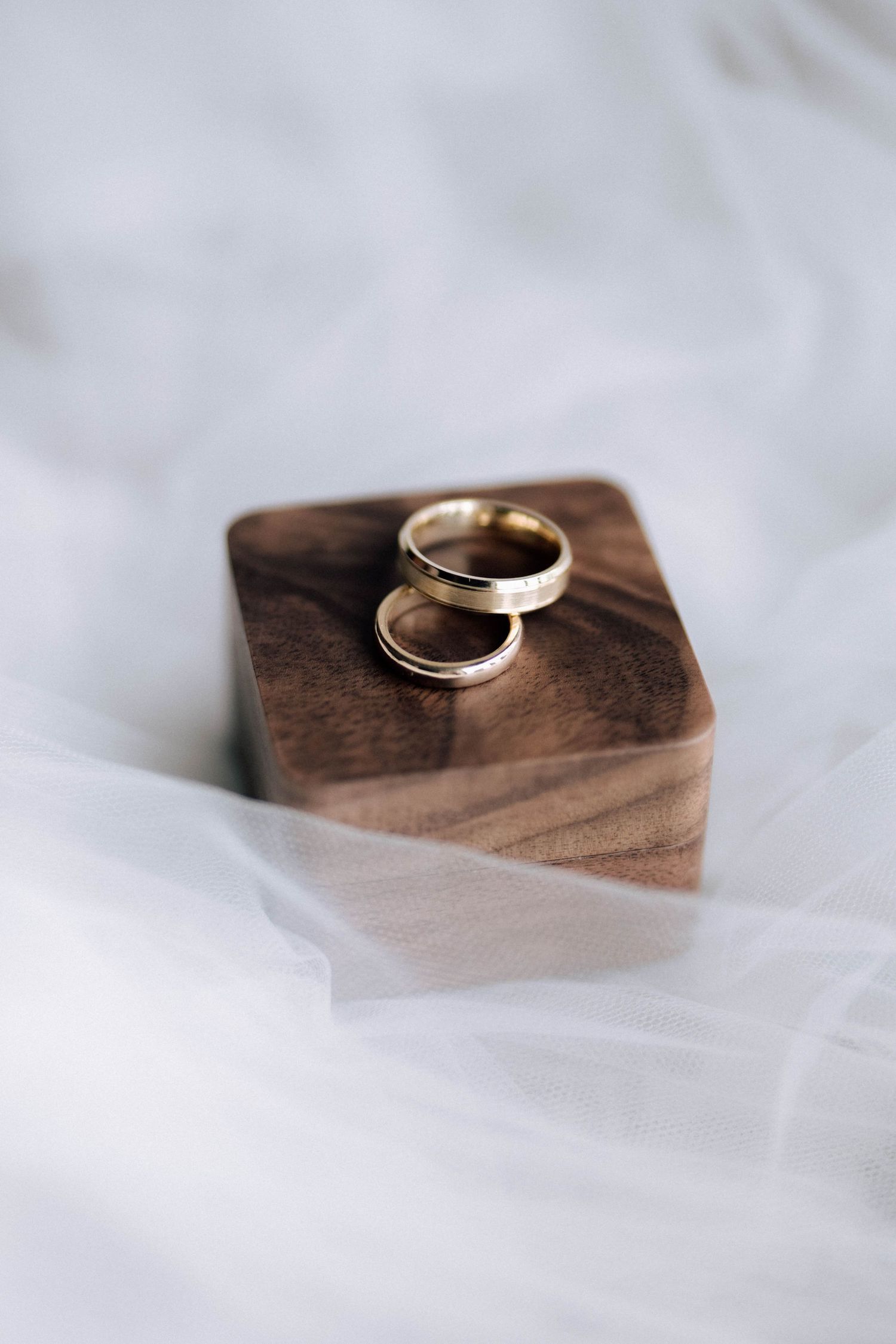Two gold wedding bands resting elegantly on a small wooden ring box atop a white bridal veil.