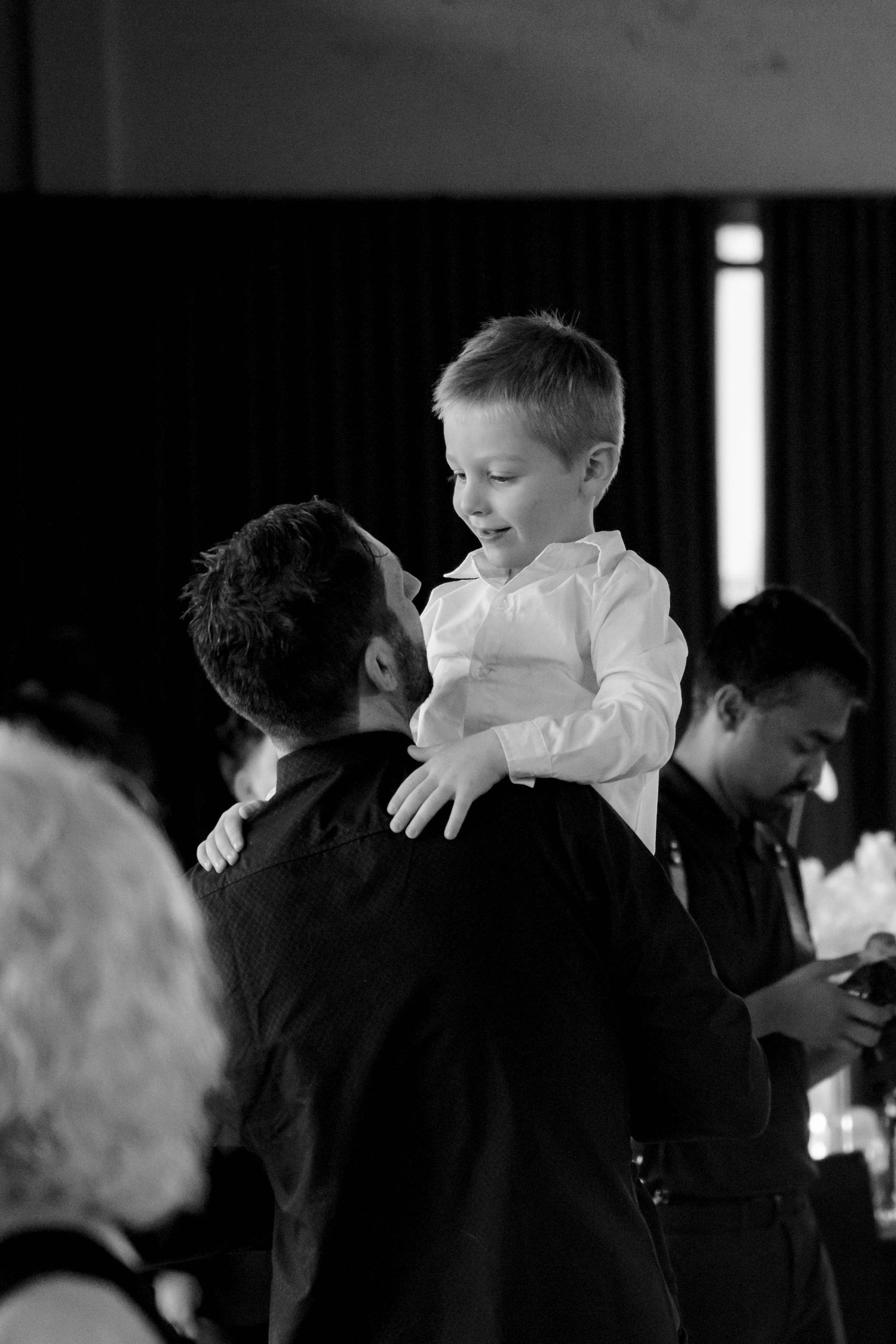 A joyful young boy in a white shirt being lifted up by a man in black at a wedding reception.