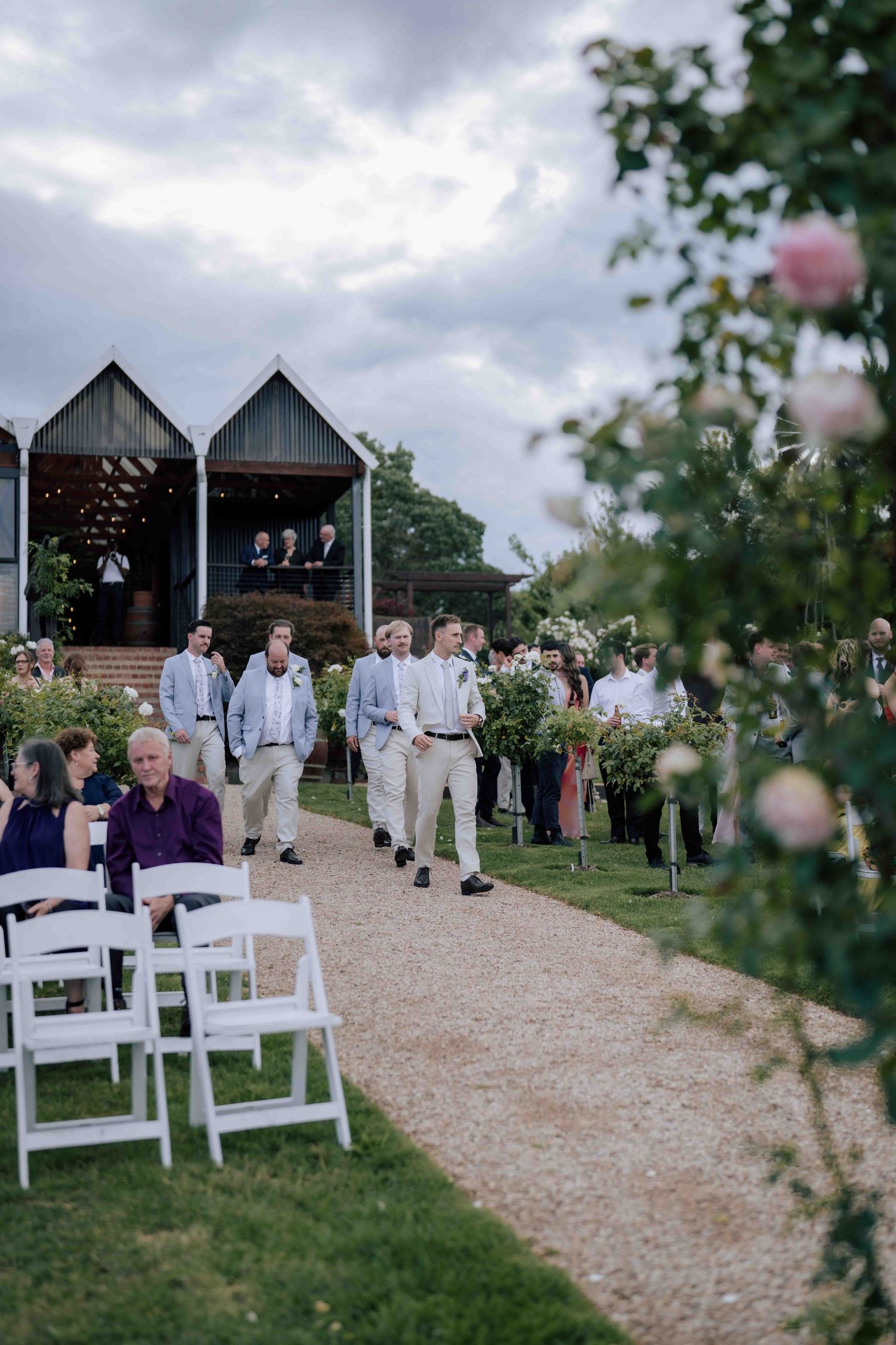 Groomsmen walk down outdoor wedding aisle lined with white chairs and pink roses at a barn venue.