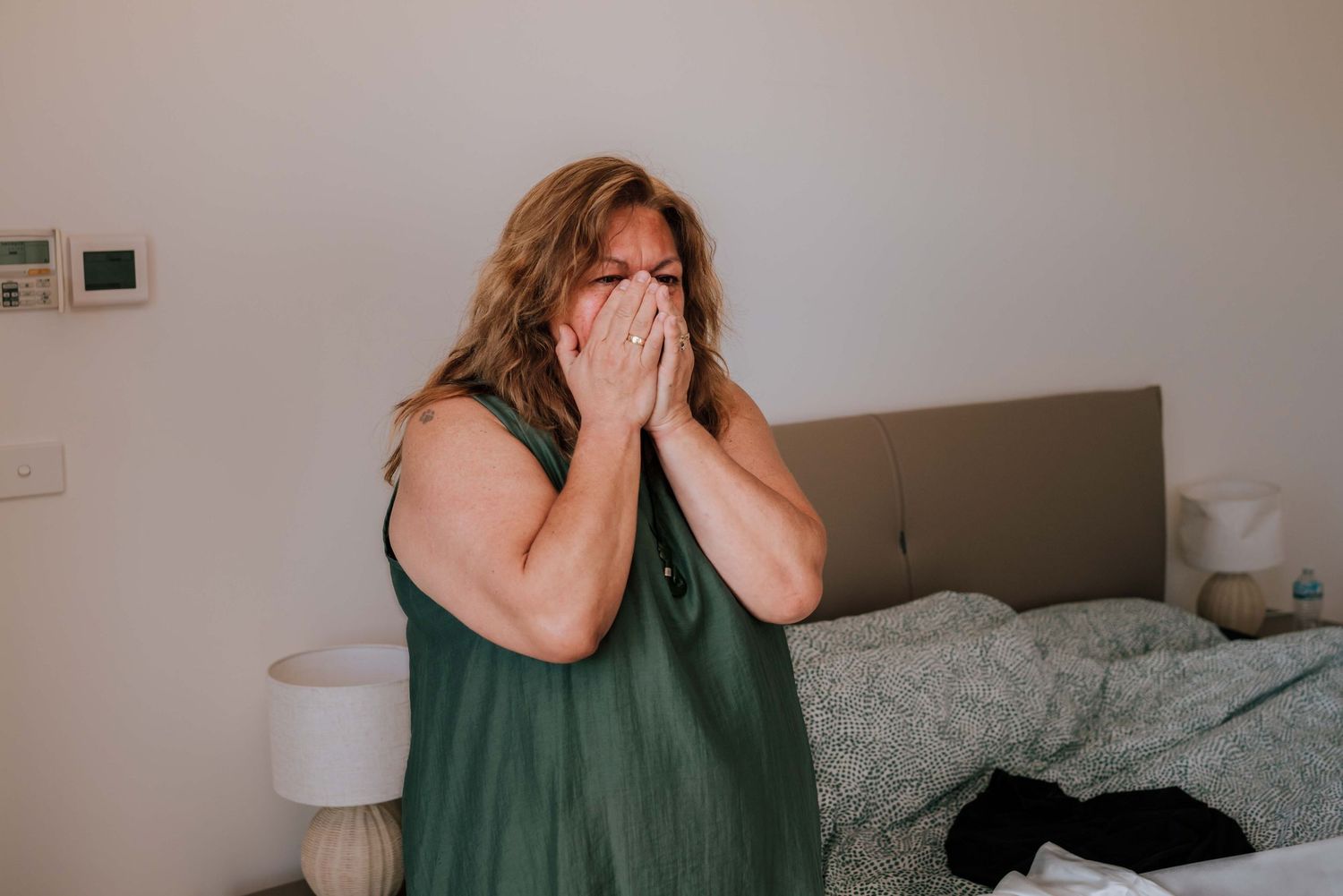 Emotional woman in green dress covering face with both hands while sitting on edge of bed indoors.