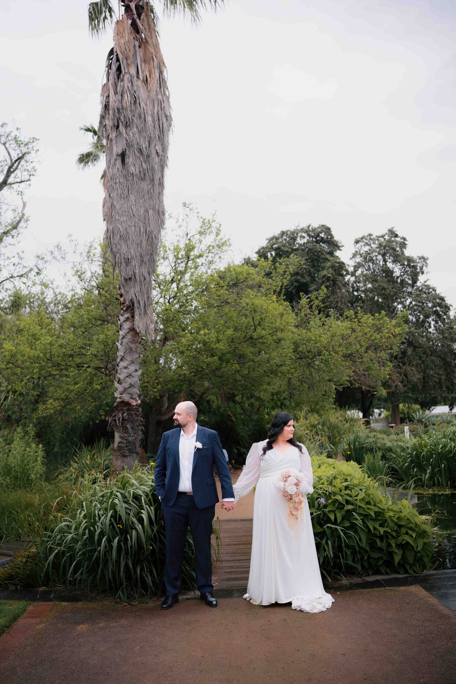 Bride and groom hold hands walking through a lush garden with tall palm tree on their wedding day.