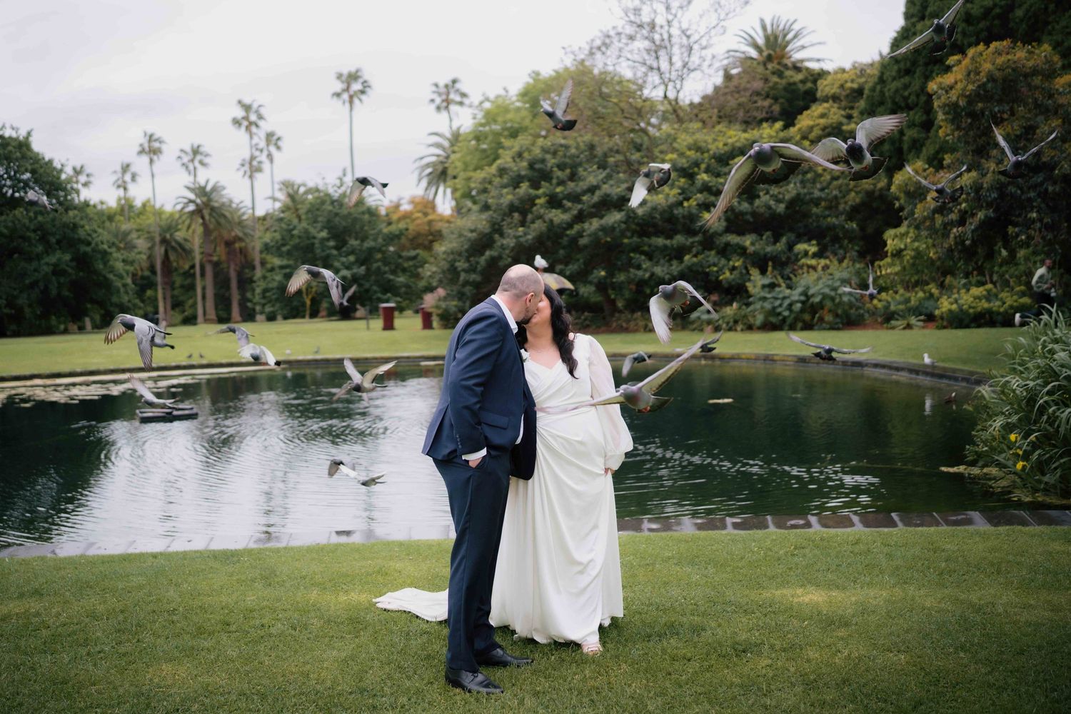 Bride and groom kissing by a pond with birds flying around them in a botanical garden.