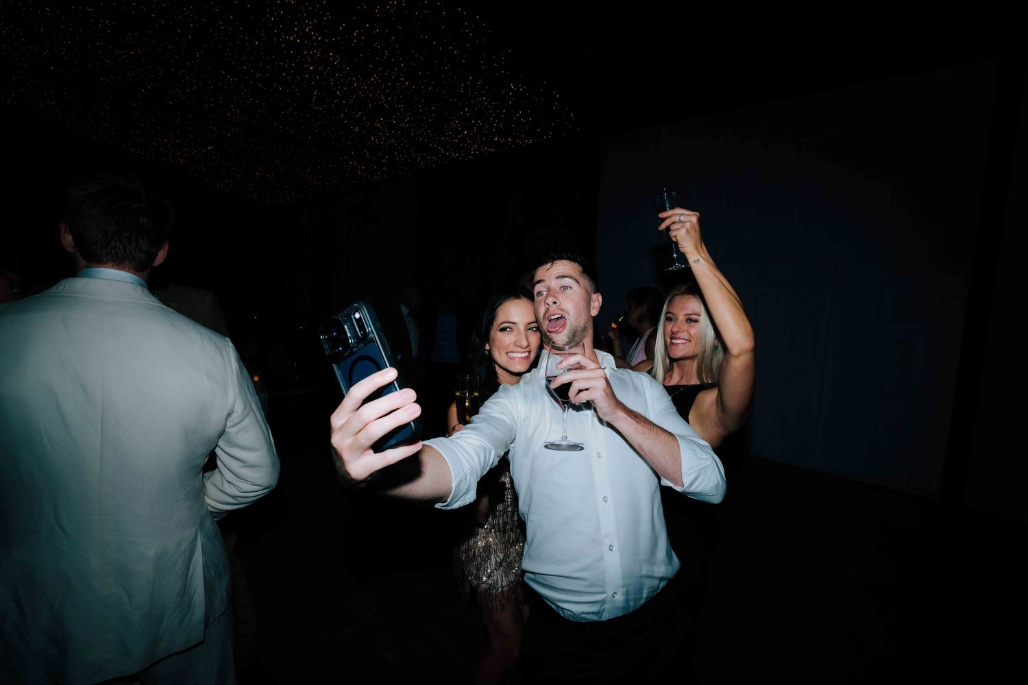 Man taking selfie with two women on dark wedding reception dance floor.