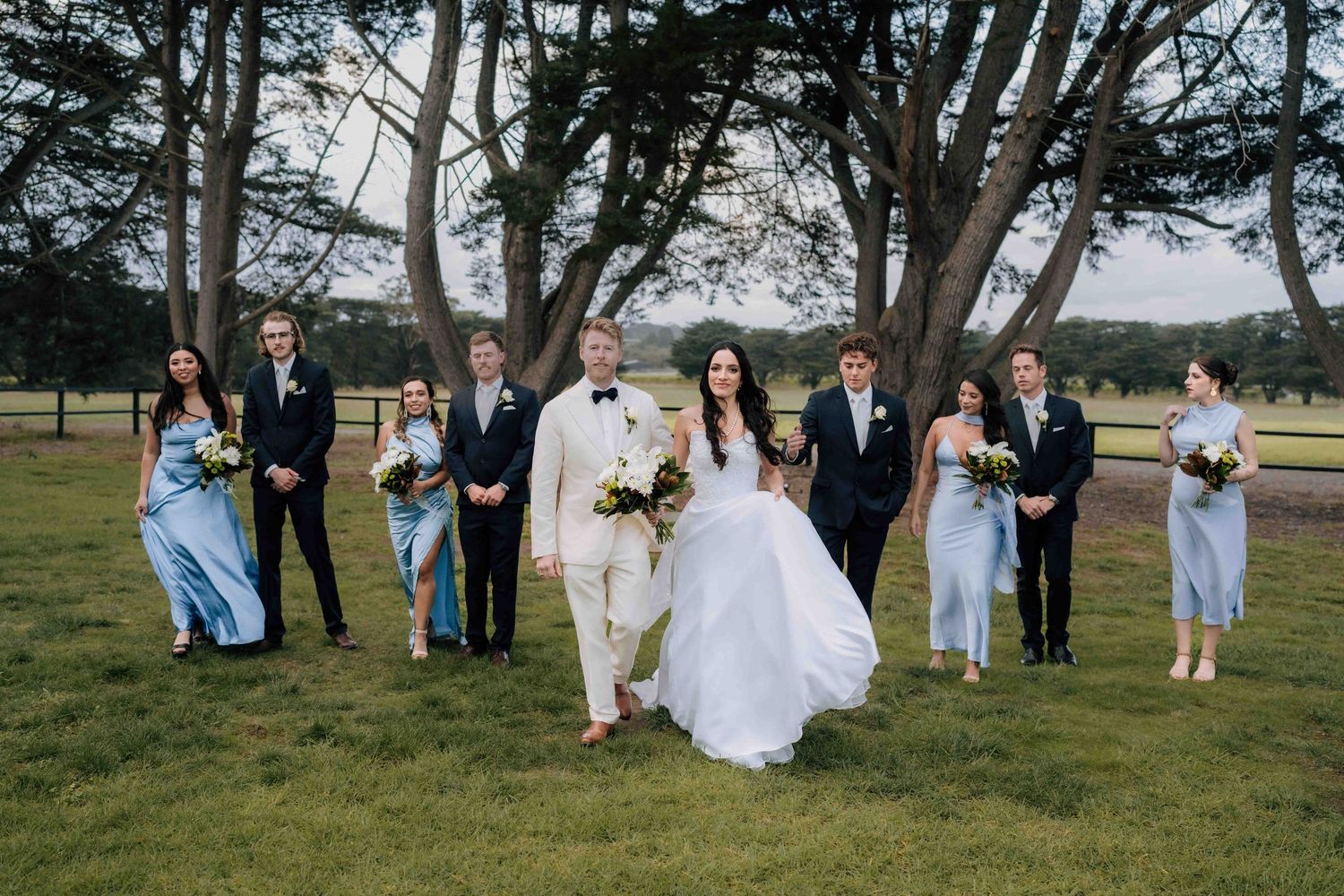 Wedding party walks outdoors on lush green lawn, bride in white gown, bridesmaids in blue dresses, groomsmen in black suits.