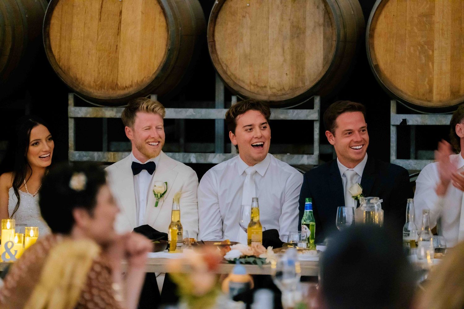 Groom in white tuxedo laughs with groomsmen at wedding reception table in front of oak wine barrels.
