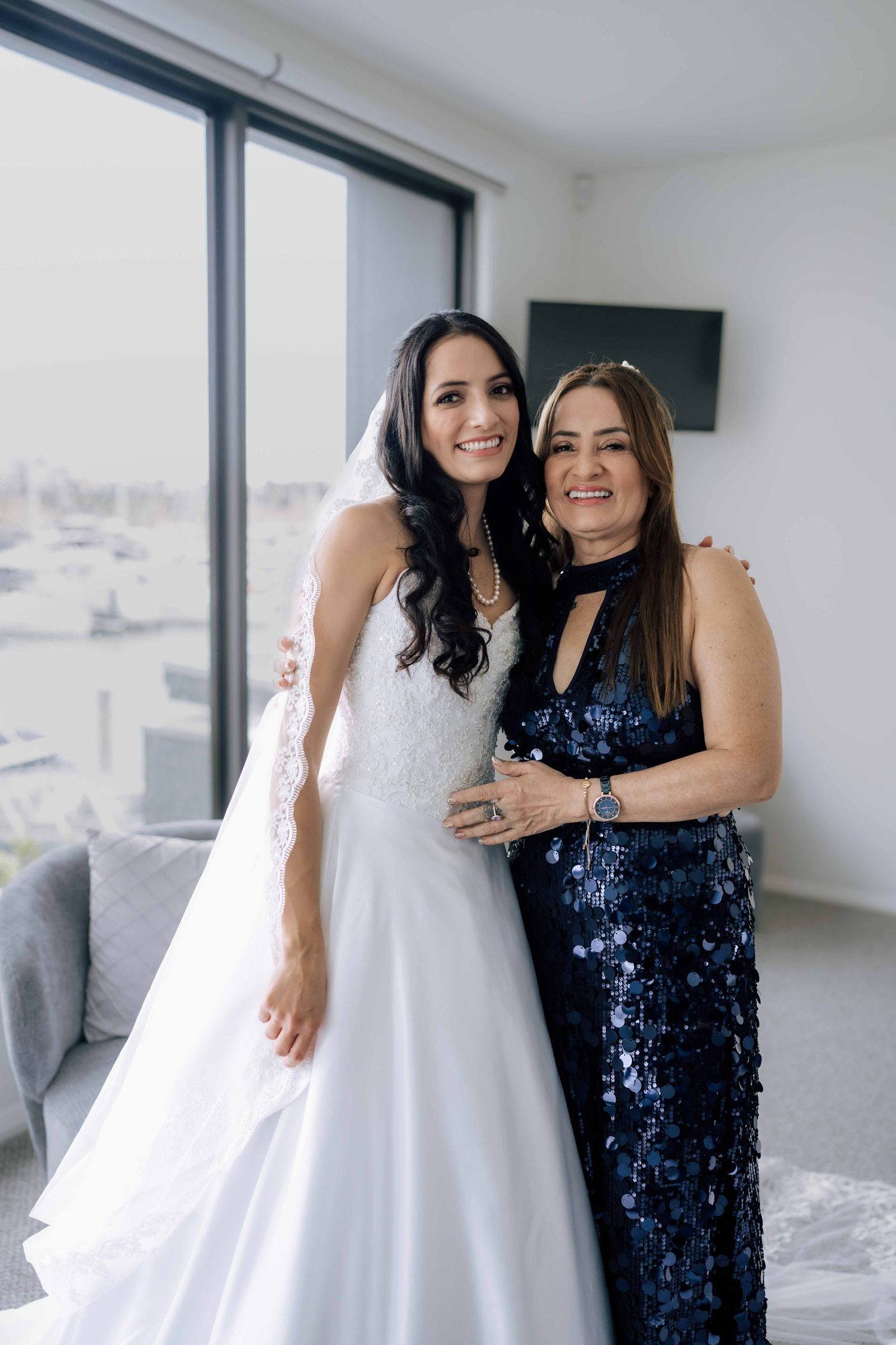 Smiling bride in white wedding dress embraces older woman in navy sequin dress by window.