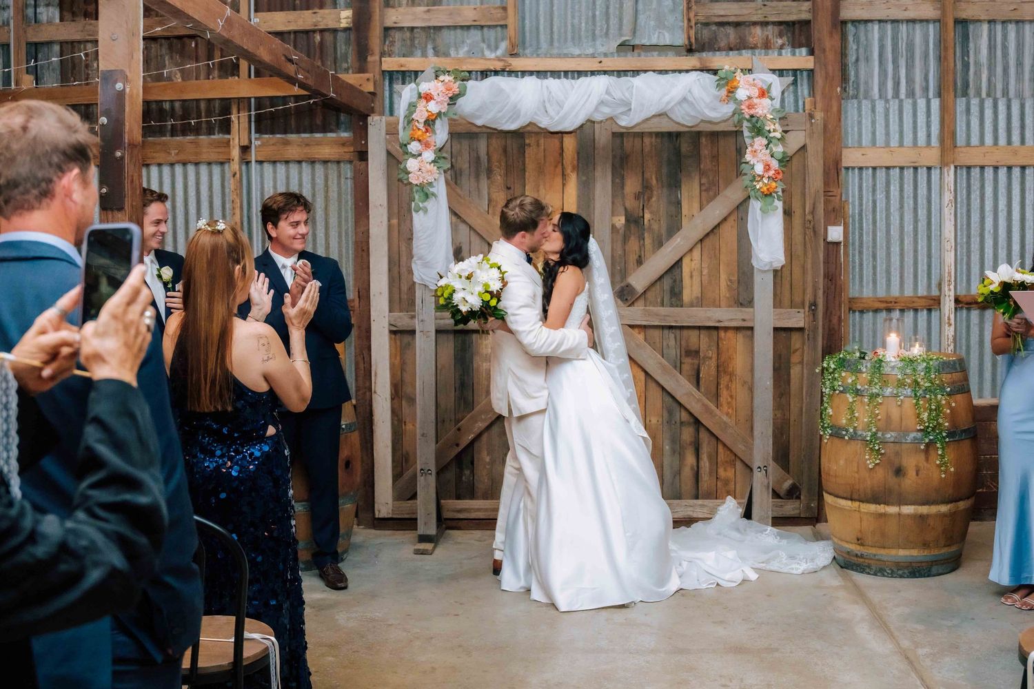 Bride and groom share first kiss at rustic barn wedding ceremony with floral arch and cheering guests.