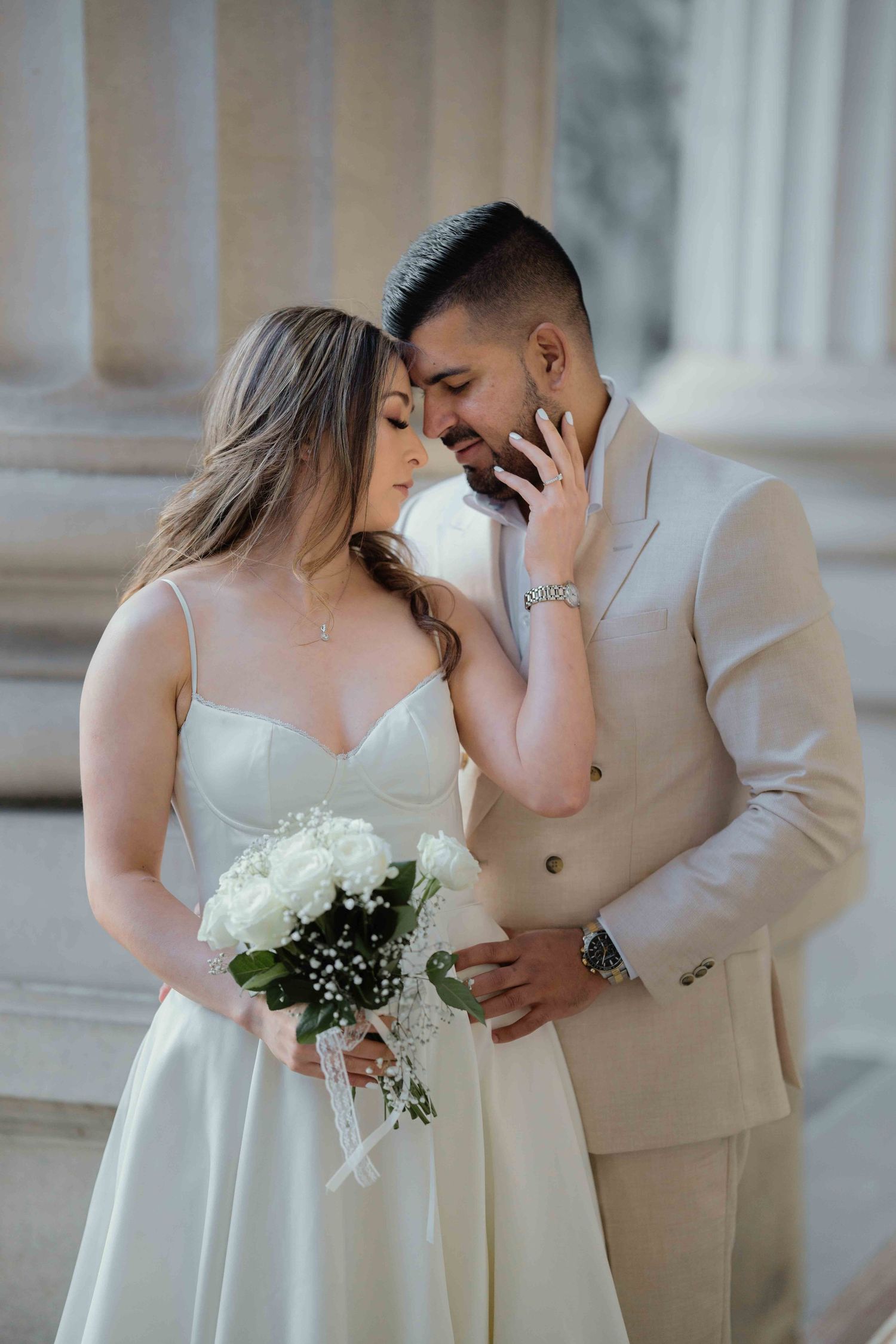 Romantic wedding portrait of a bride holding white roses bouquet leaning into her groom beside stone columns.