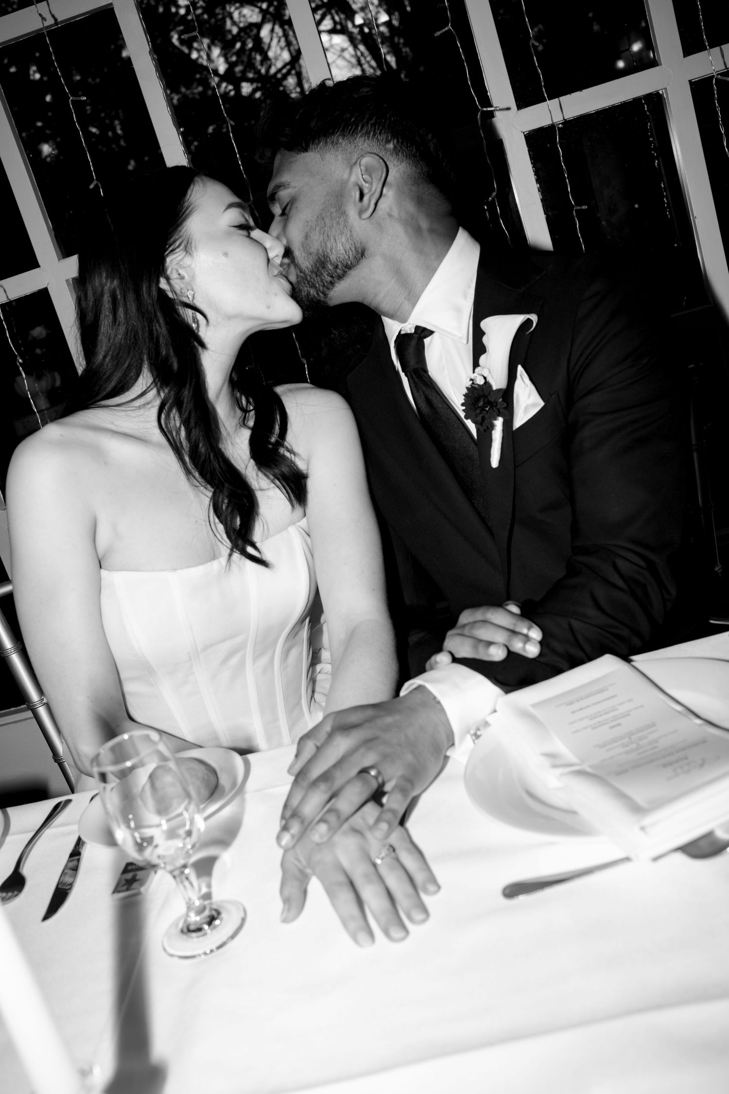 Black and white photo of a bride and groom sharing a kiss at their wedding reception table, rings visible.