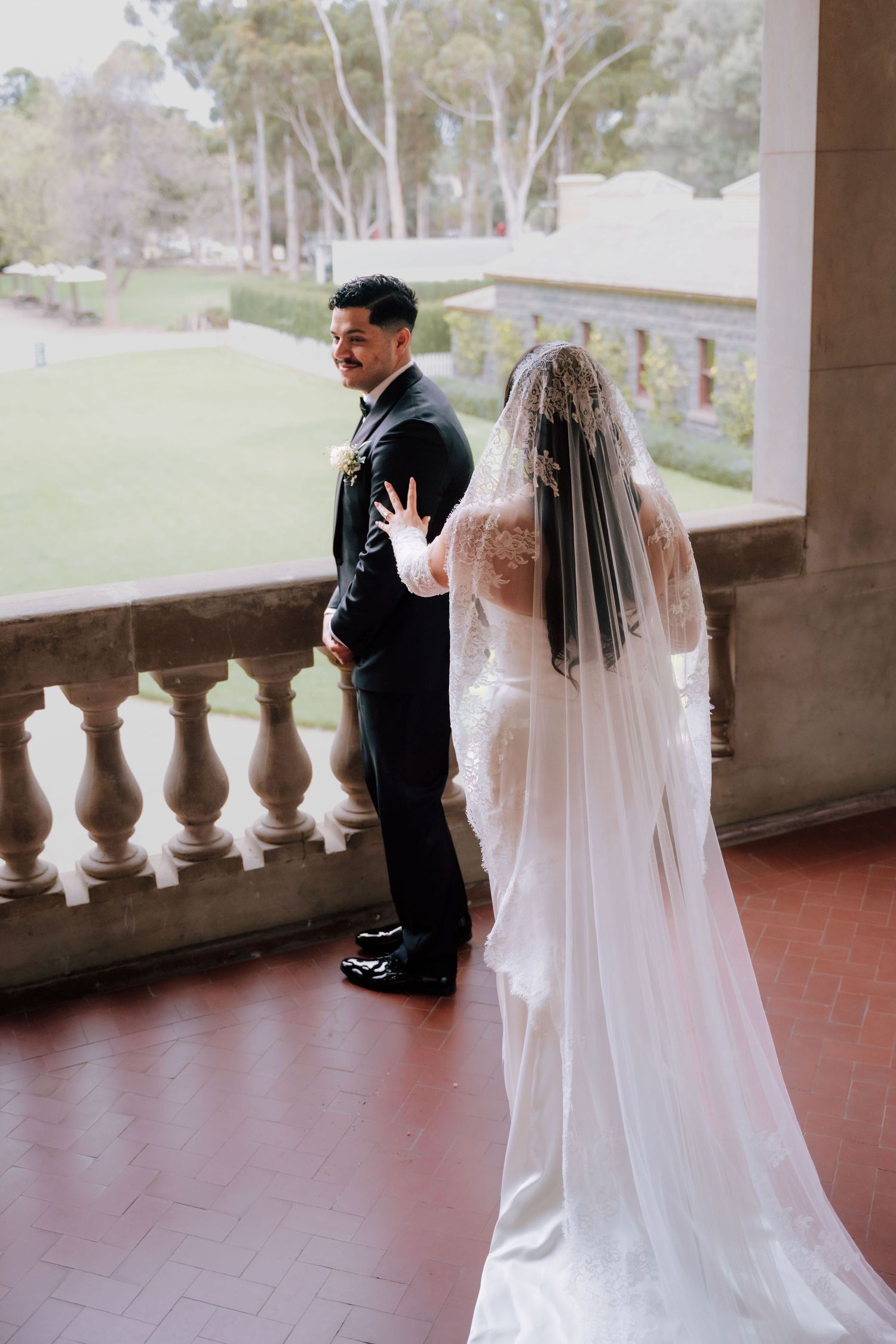 Bride in a lace veil tapping groom on shoulder during wedding first look on a balcony.