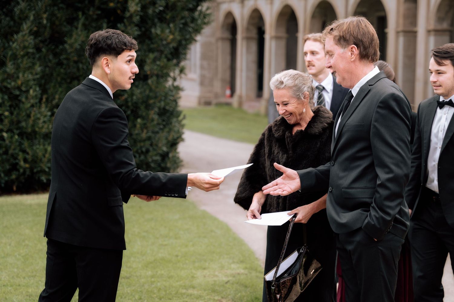 Groomsman in tuxedo handing ceremony programs to wedding guests in an outdoor courtyard setting.