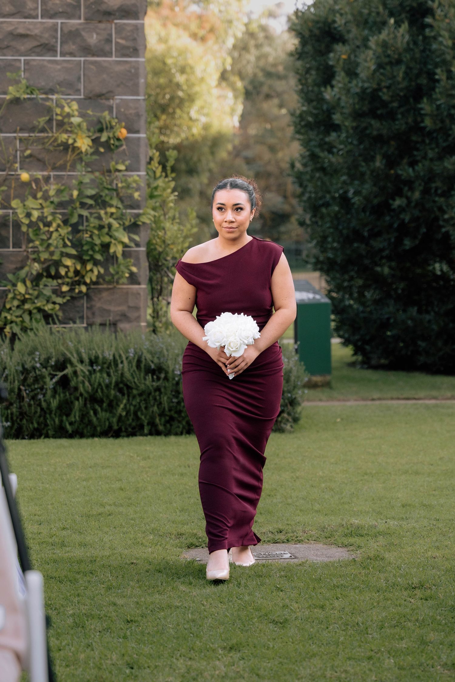 Bridesmaid in burgundy gown holding white bouquet walking across green lawn at wedding.